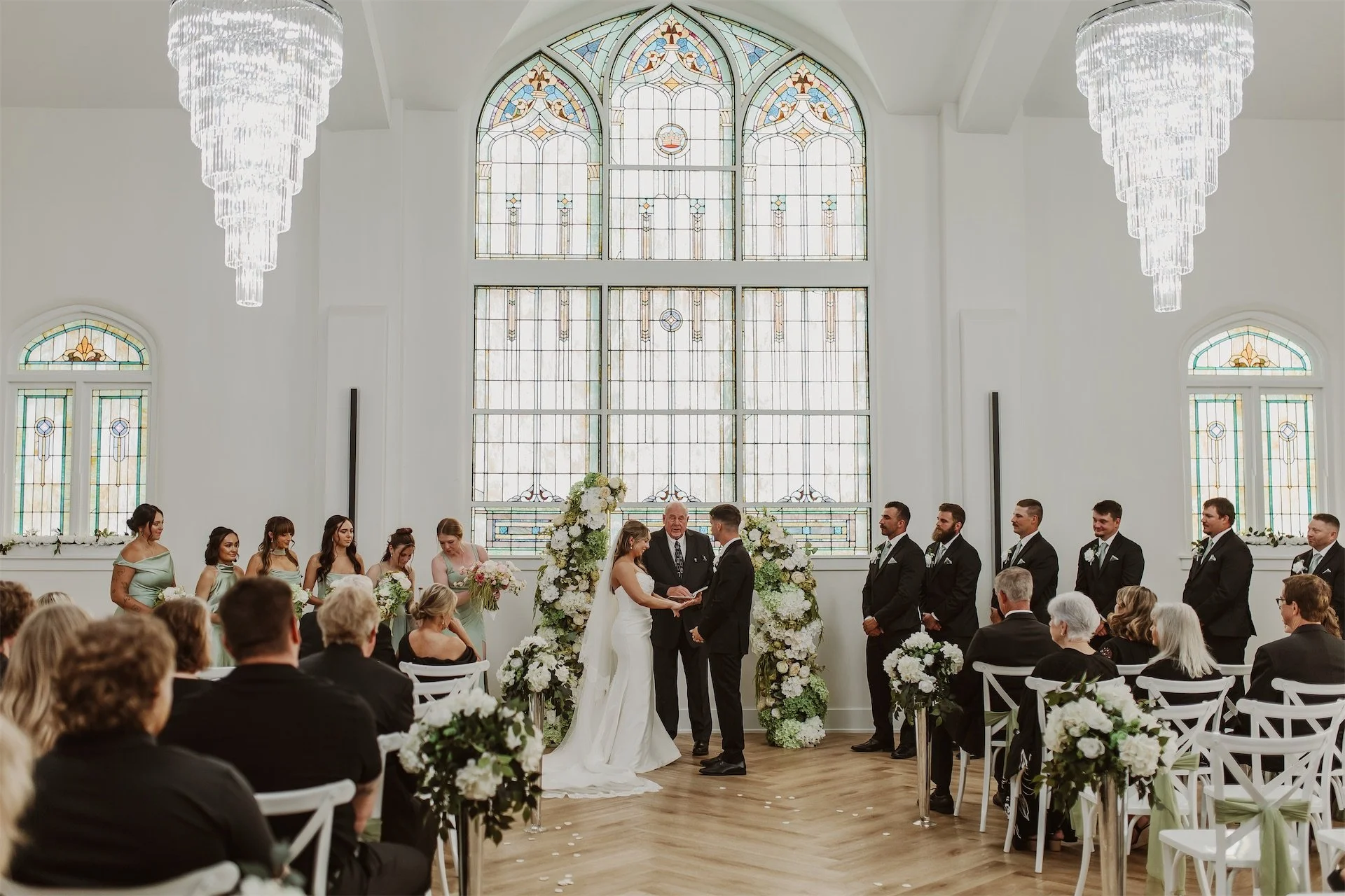Groom teary eyed listening to officiant during vows beneath stained glass window