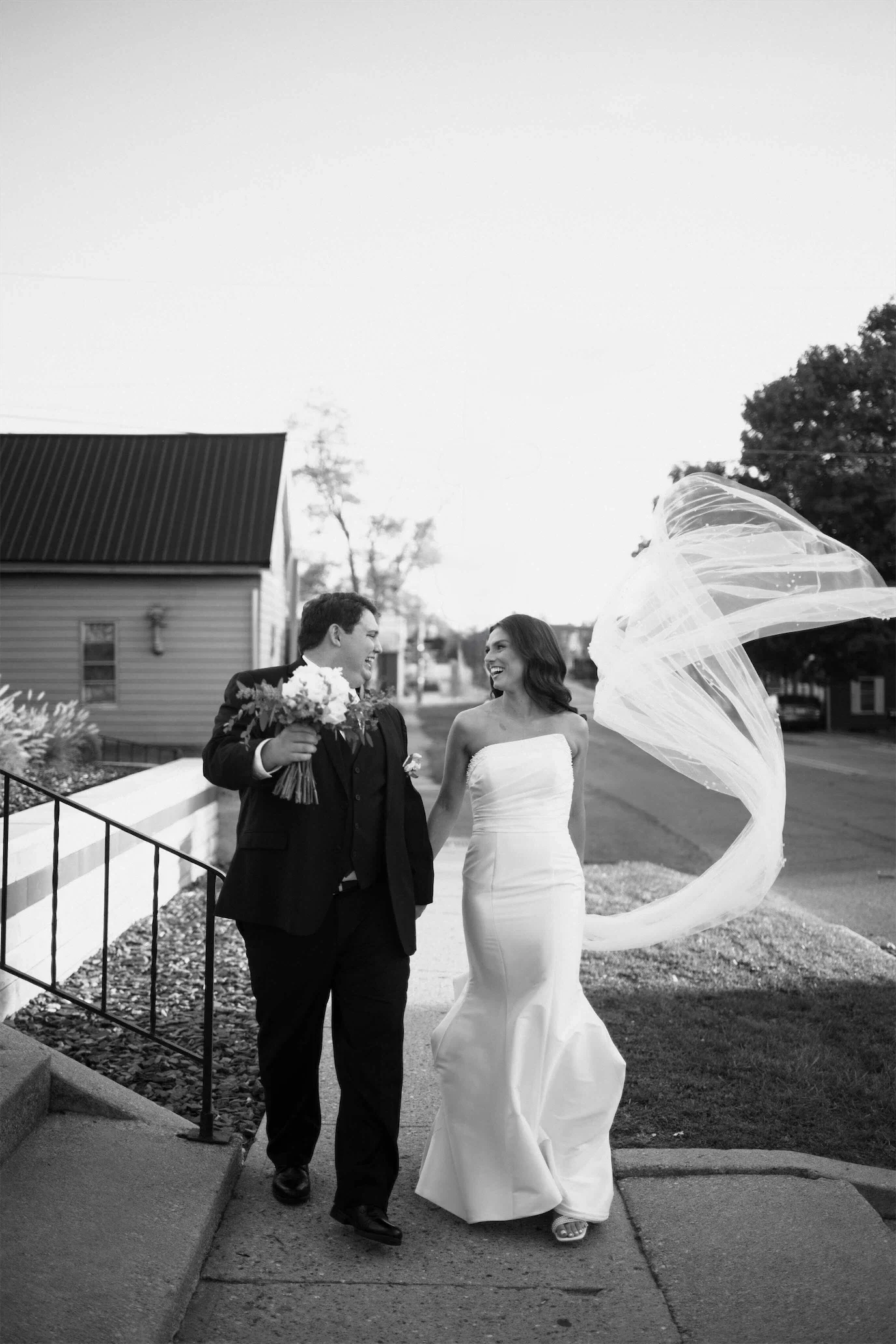 Black and white photo of the couple walking hand in hand as the bride's long veil blows in the wind