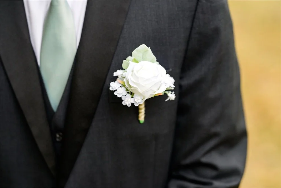 Close up of a groom's sage green tie, charcoal vest and white rose boutonniere