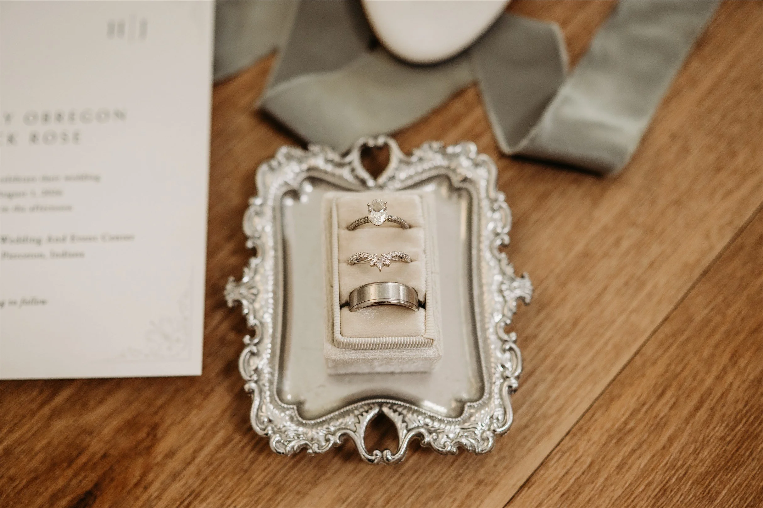 Wedding ring set in a cream velvet box on an ornate silver tray beside a sage green ribbon and invitation