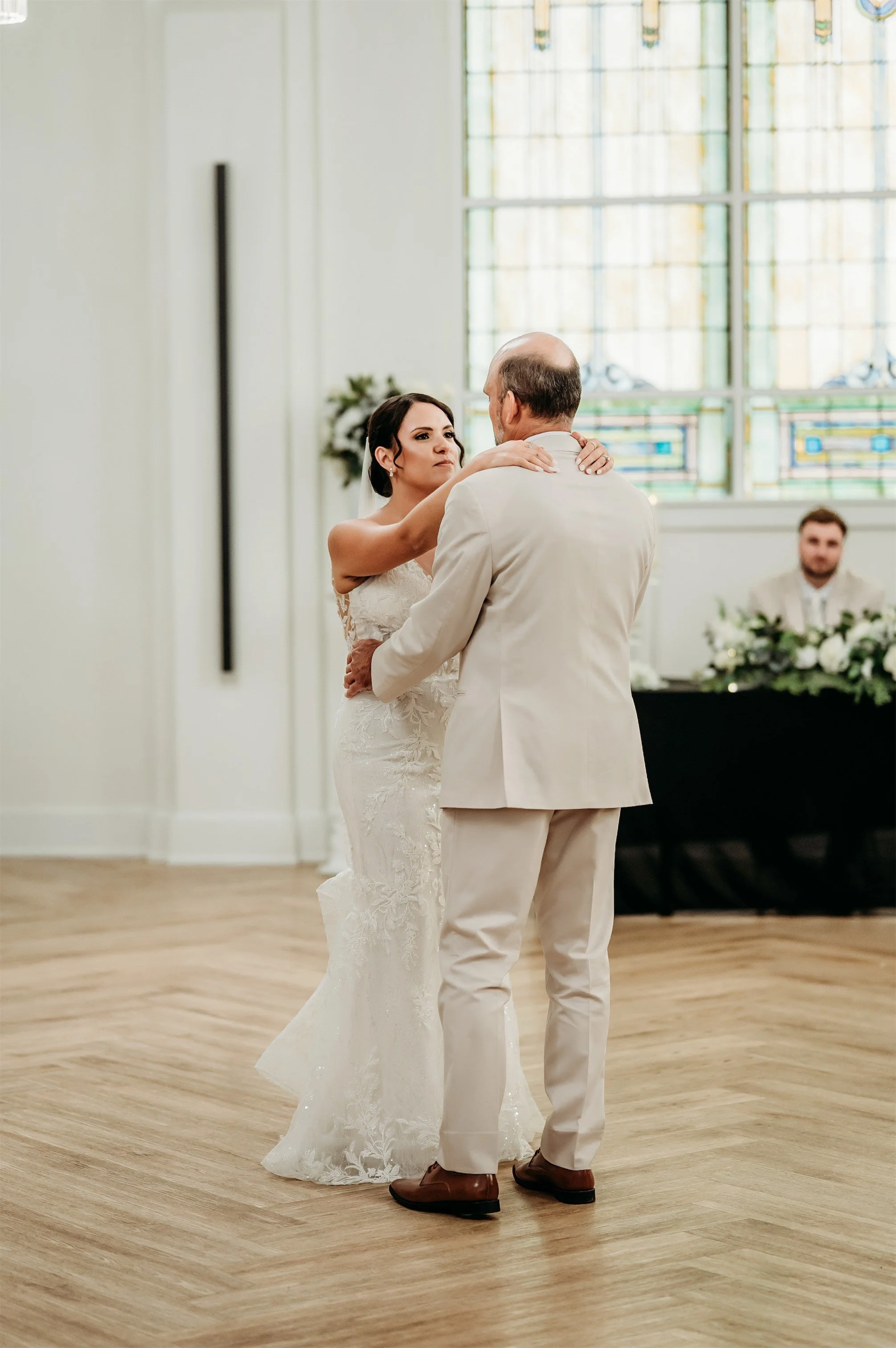 Bride dancing with her father in a cream suit on a herringbone wood floor near stained glass windows
