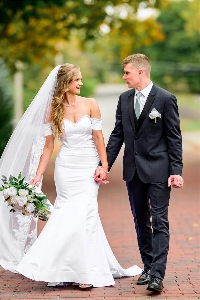 Bride and groom walk hand in hand along a brick path and smile at each other