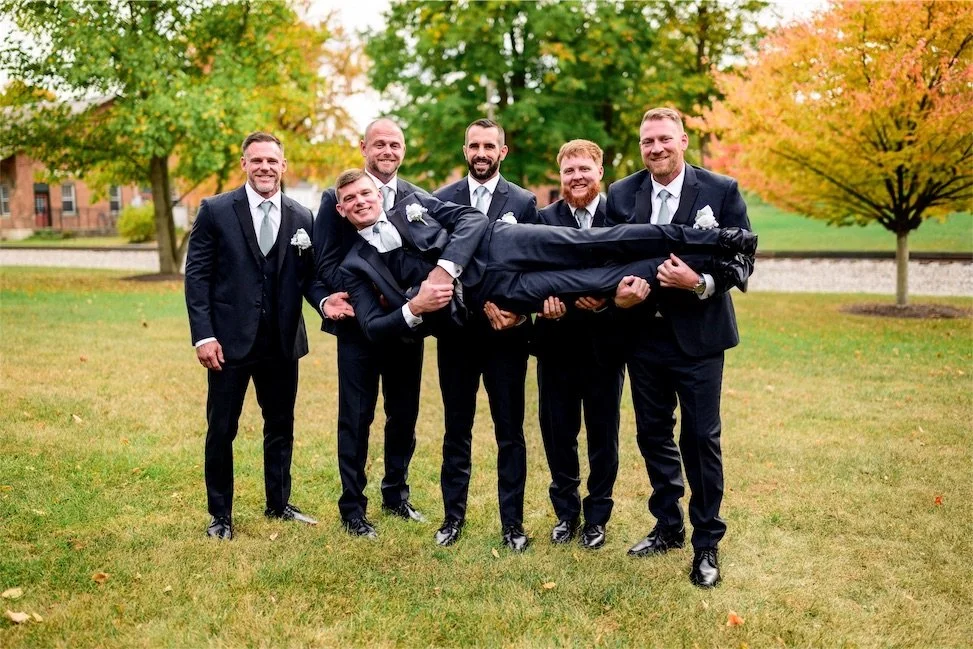 Groomsmen in black suits lift the groom horizontally while laughing on a grassy lawn in autumn