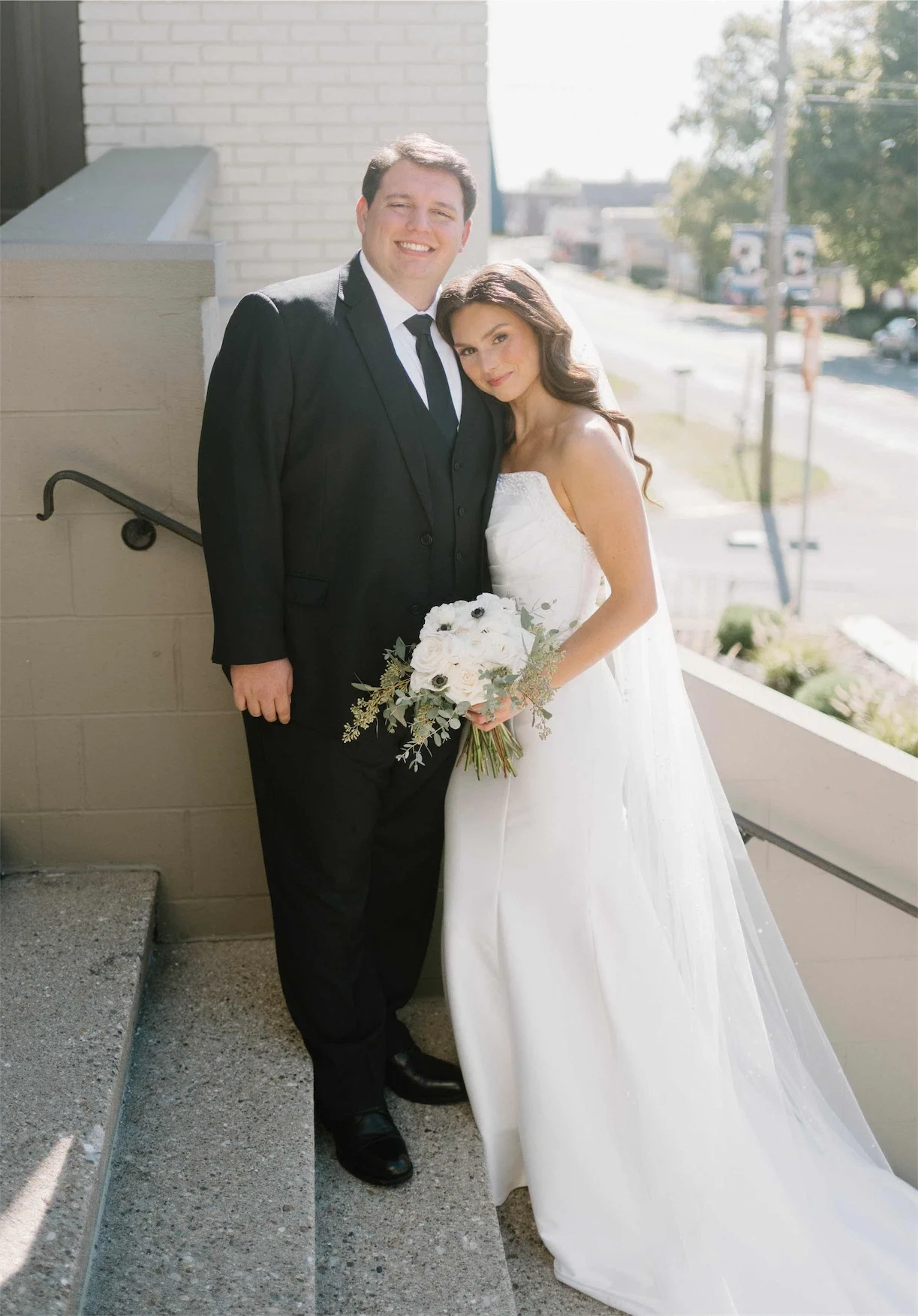 Bride and groom smiling together on the sunny front steps of a converted brick chapel