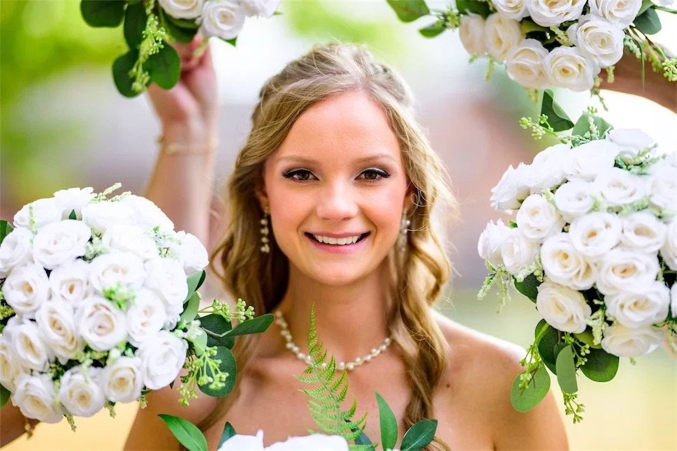 Smiling bride in a pearl necklace framed by white rose bouquets held by her bridesmaids