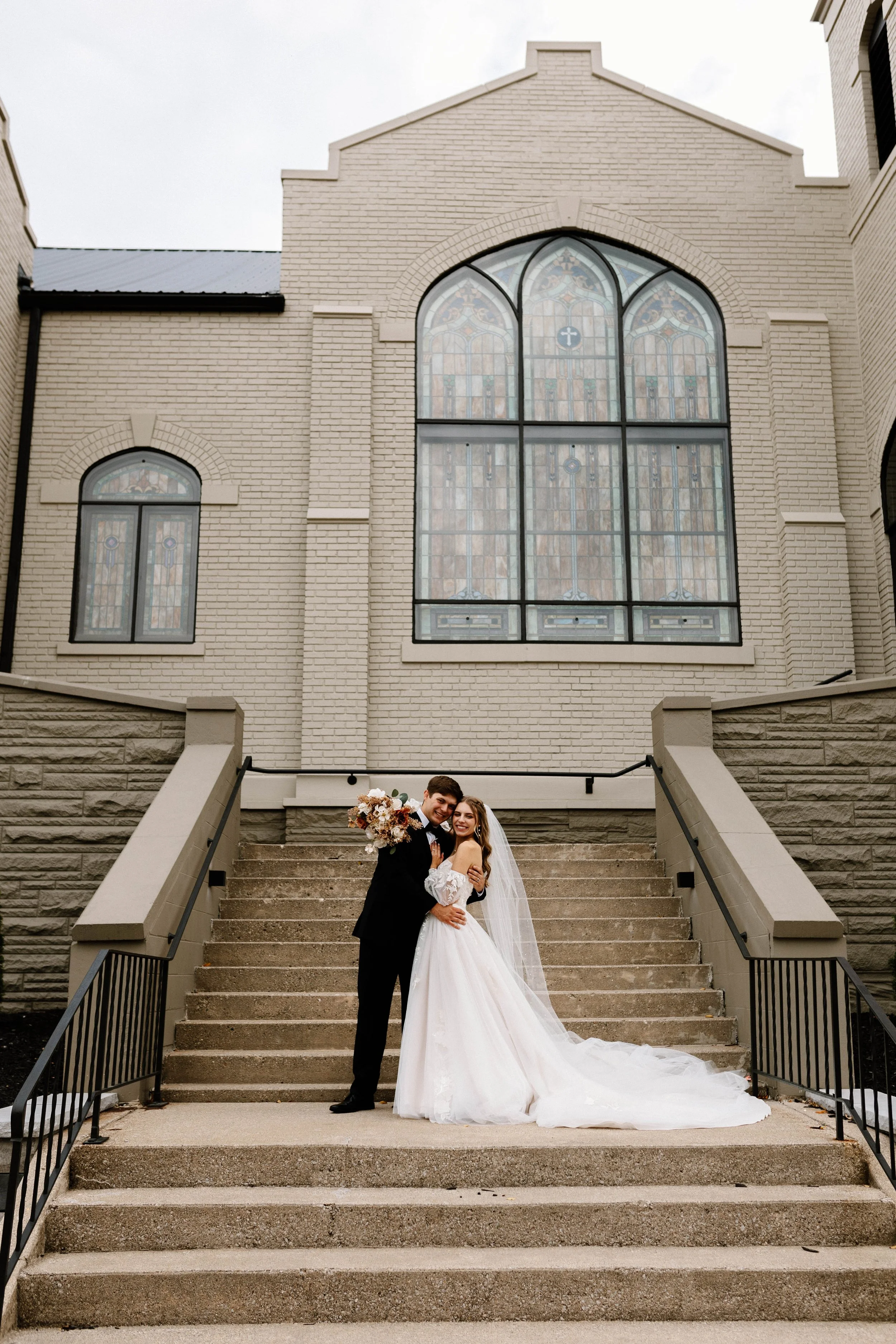 A bride and groom in wedding attire standing on a staircase in front of a church with large stained glass windows. The bride is holding a bouquet of flowers and wearing a long white gown with a veil. The groom is in a black suit, and they are smiling