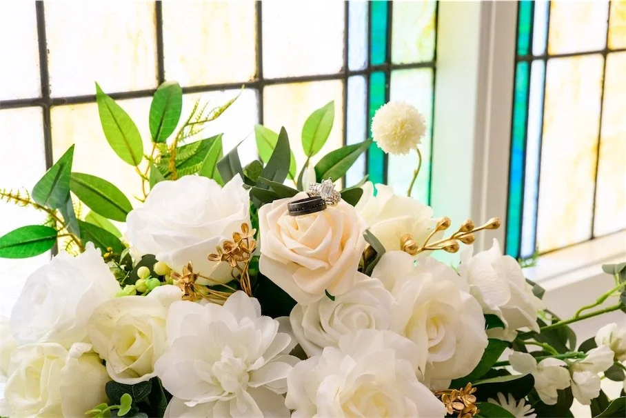 Wedding bands resting in a white rose and greenery bouquet beside a stained glass window