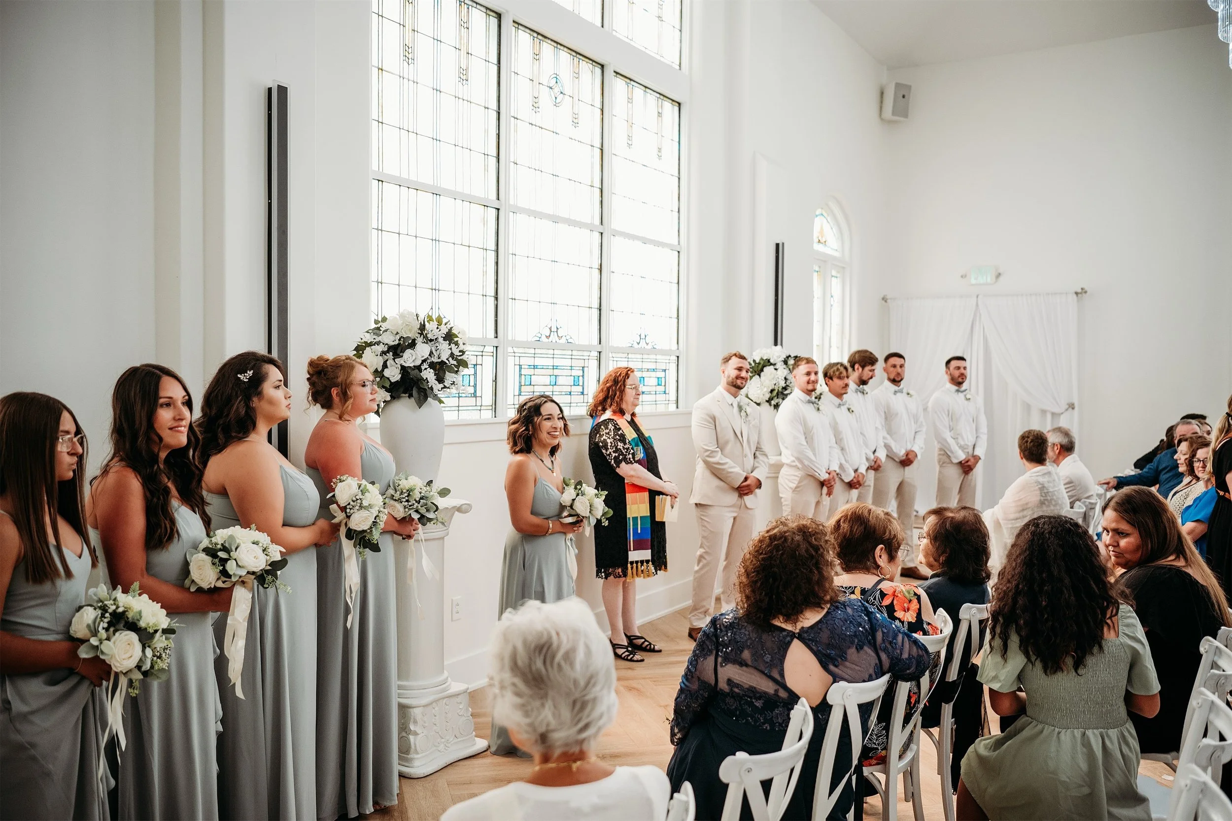 Bridesmaids in sage green gowns and groomsmen in tan suits lined up during a wedding ceremony with guests seated
