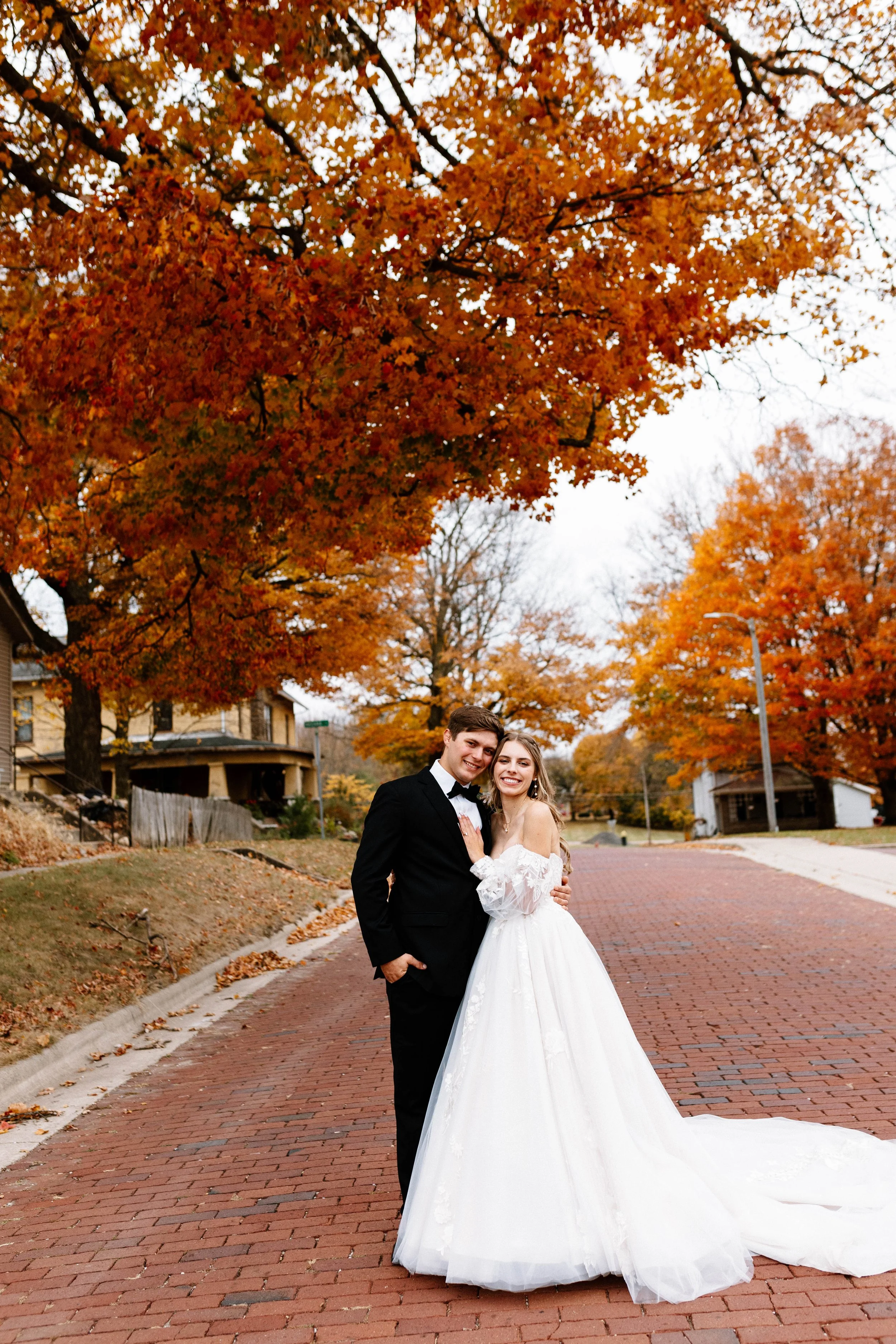A couple in wedding attire standing on a brick road in a neighborhood with fall trees with orange leaves.