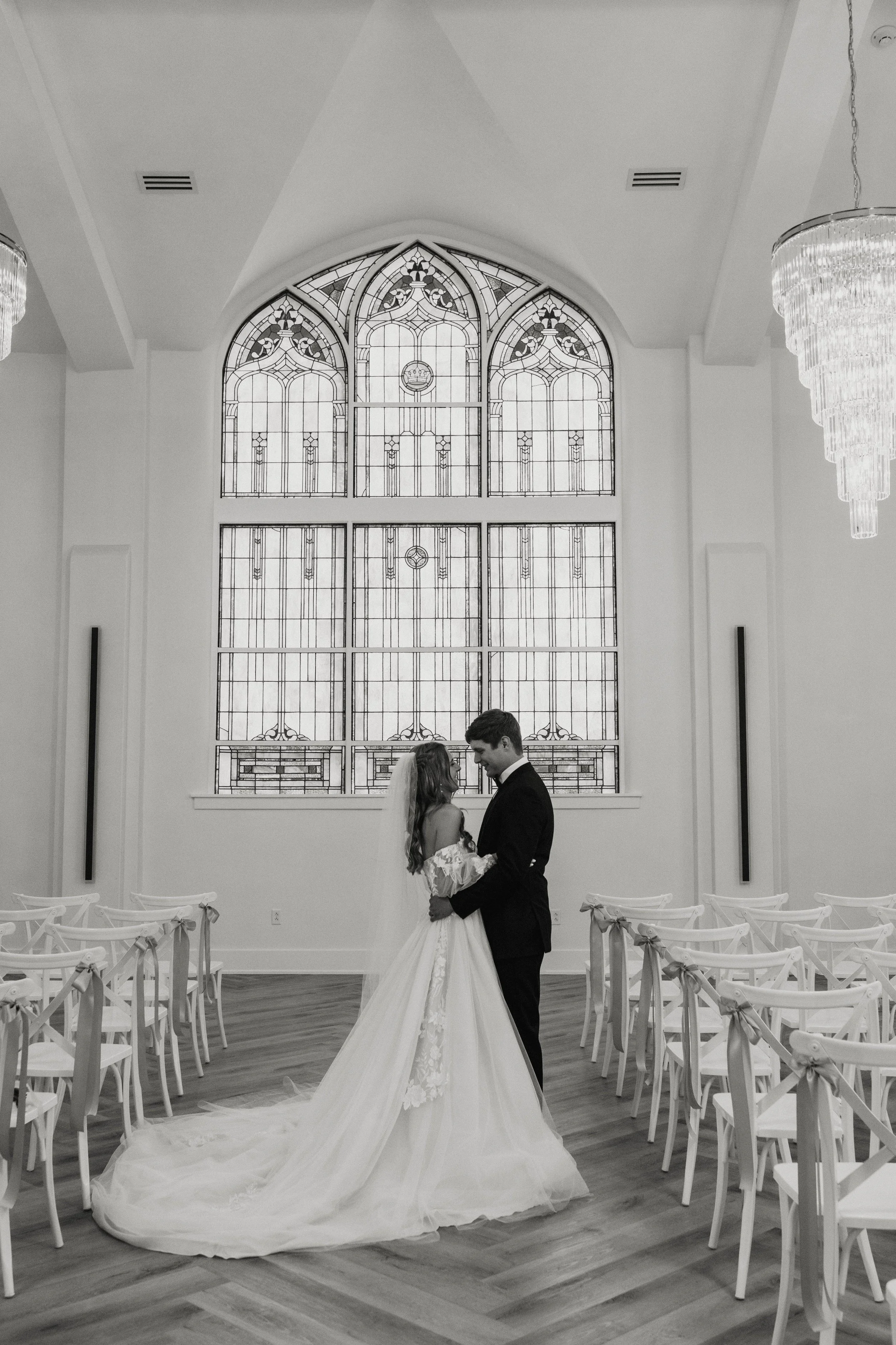 Bride and groom sharing a dance in a church with stained glass windows, surrounded by decorated chairs.