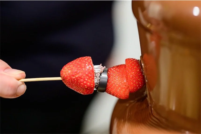 Hand dipping a strawberry threaded with a wedding band into a flowing chocolate fountain