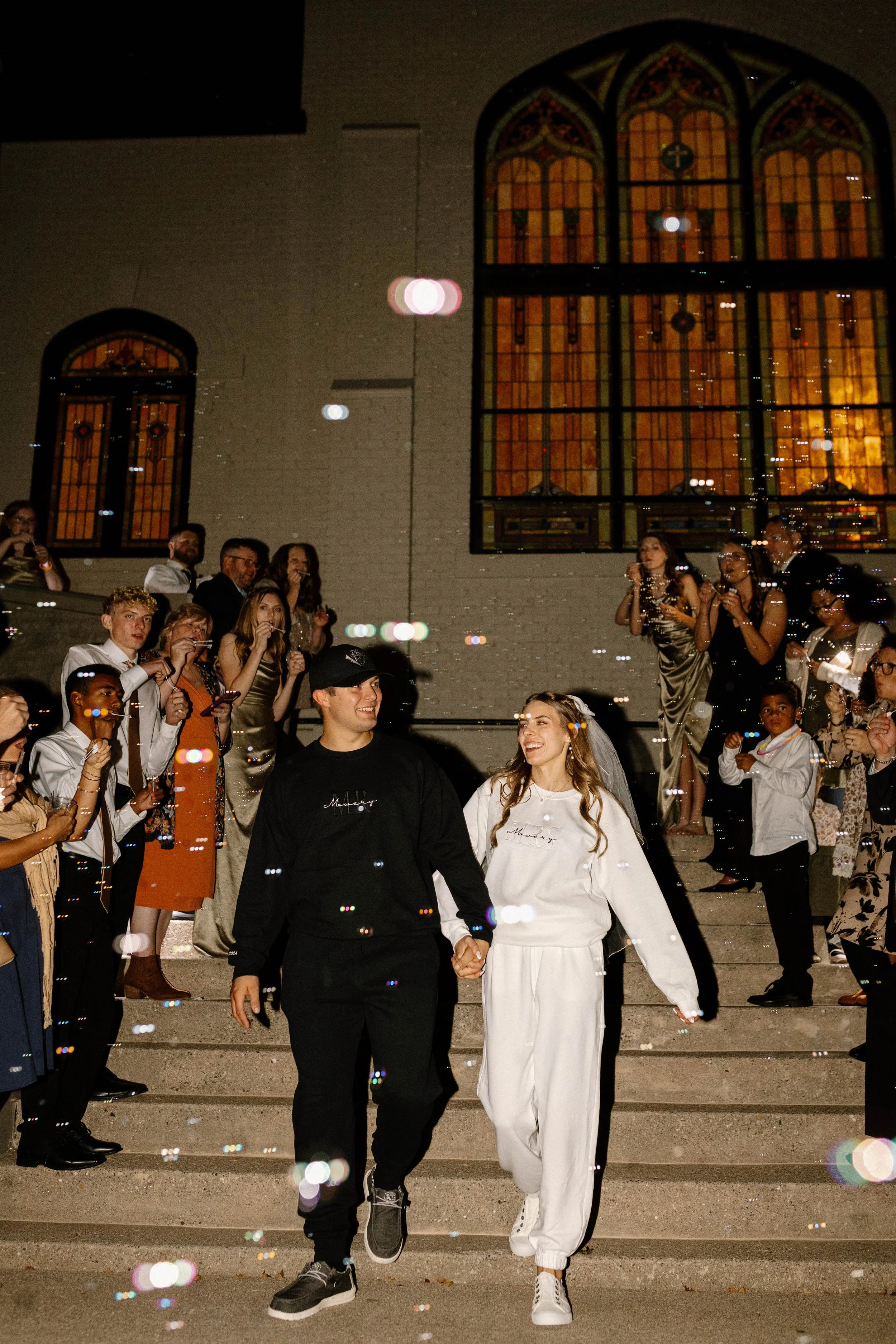 A bride and groom holding hands and walking down the stairs outside a church, surrounded by friends and family celebrating, at night with stained glass windows in the background.