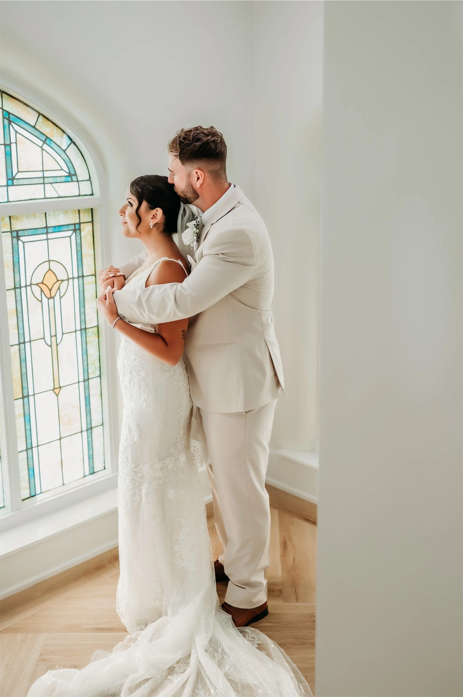 Groom kissing the bride's head as they gaze out an arched stained glass window with her train on the floor