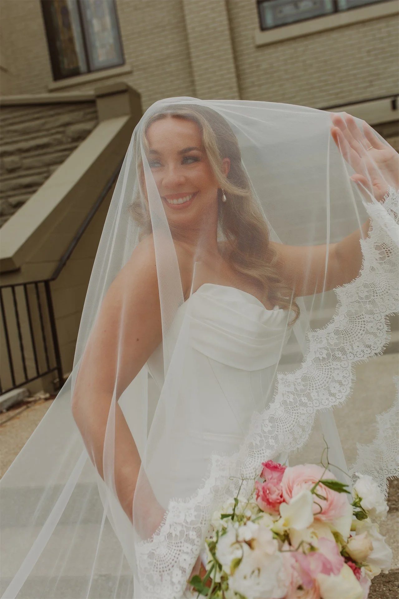 Close up bride smiling beneath veil outside stained glass church exterior