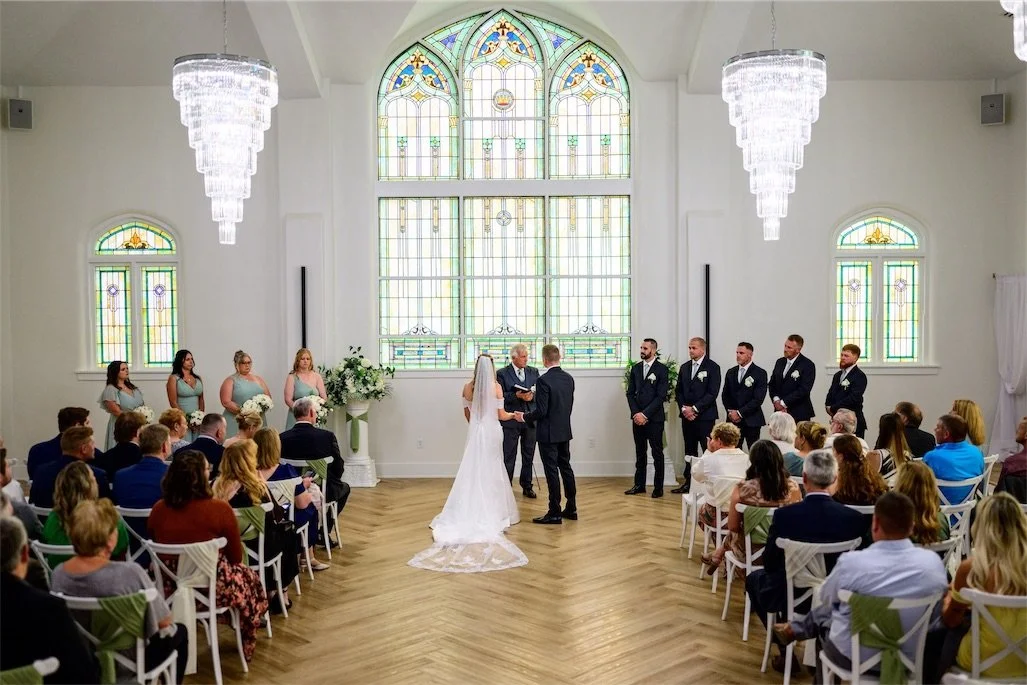 Wide view of a wedding ceremony beneath crystal chandeliers and a tall arched stained glass window