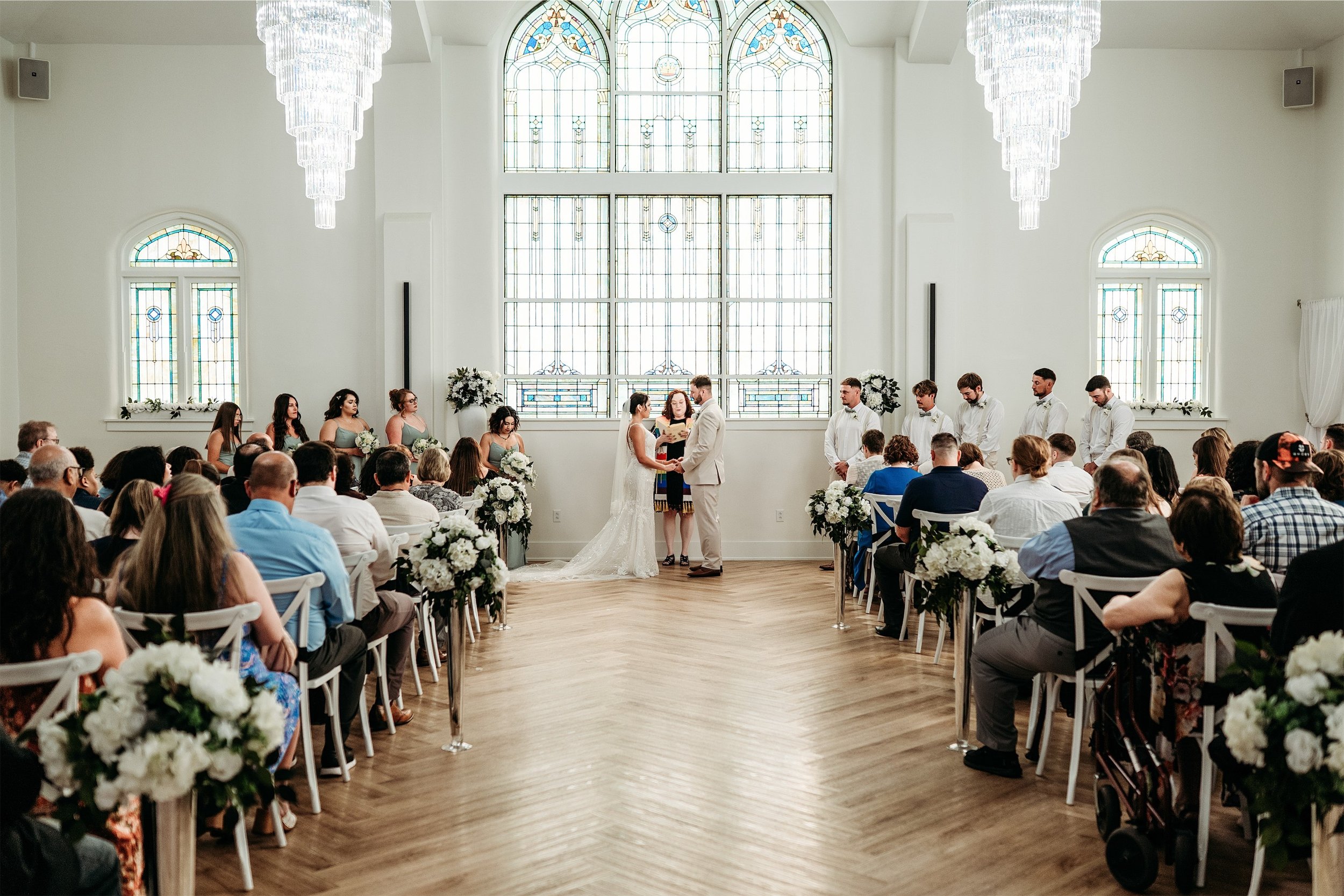Wide view of a wedding ceremony with guests seated along a herringbone aisle under two crystal chandeliers