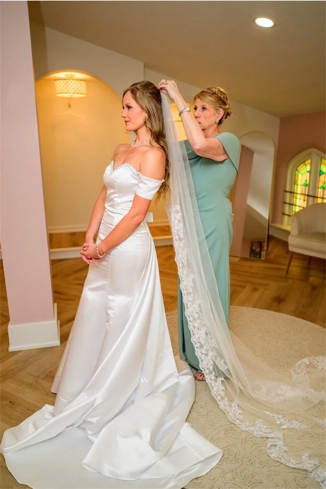Woman in a sage dress adjusts the bride's long lace veil in the bridal suite
