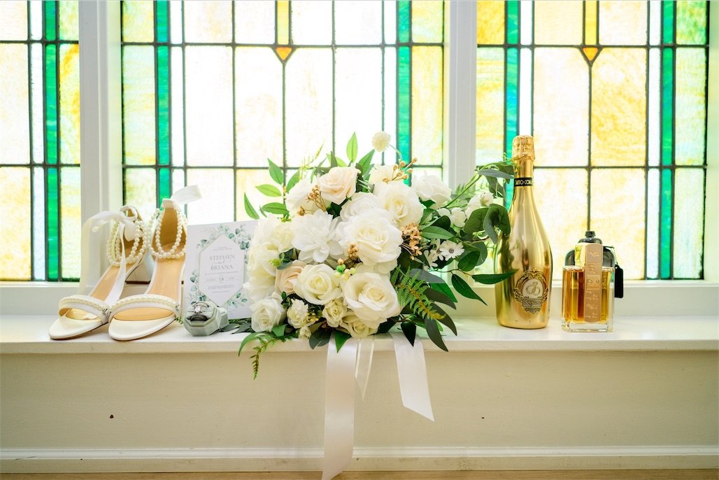 Gold champagne bottle, perfume, pearl heels and a white rose bouquet lined up before stained glass windows