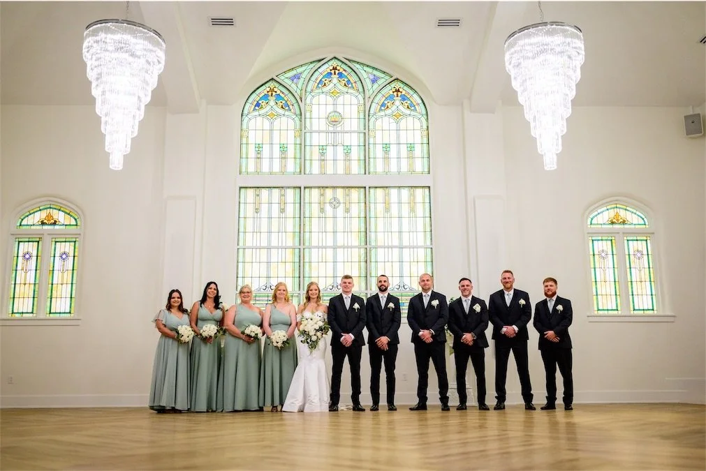 Bridesmaids in sage green and groomsmen in black suits stand in a line beneath chandeliers