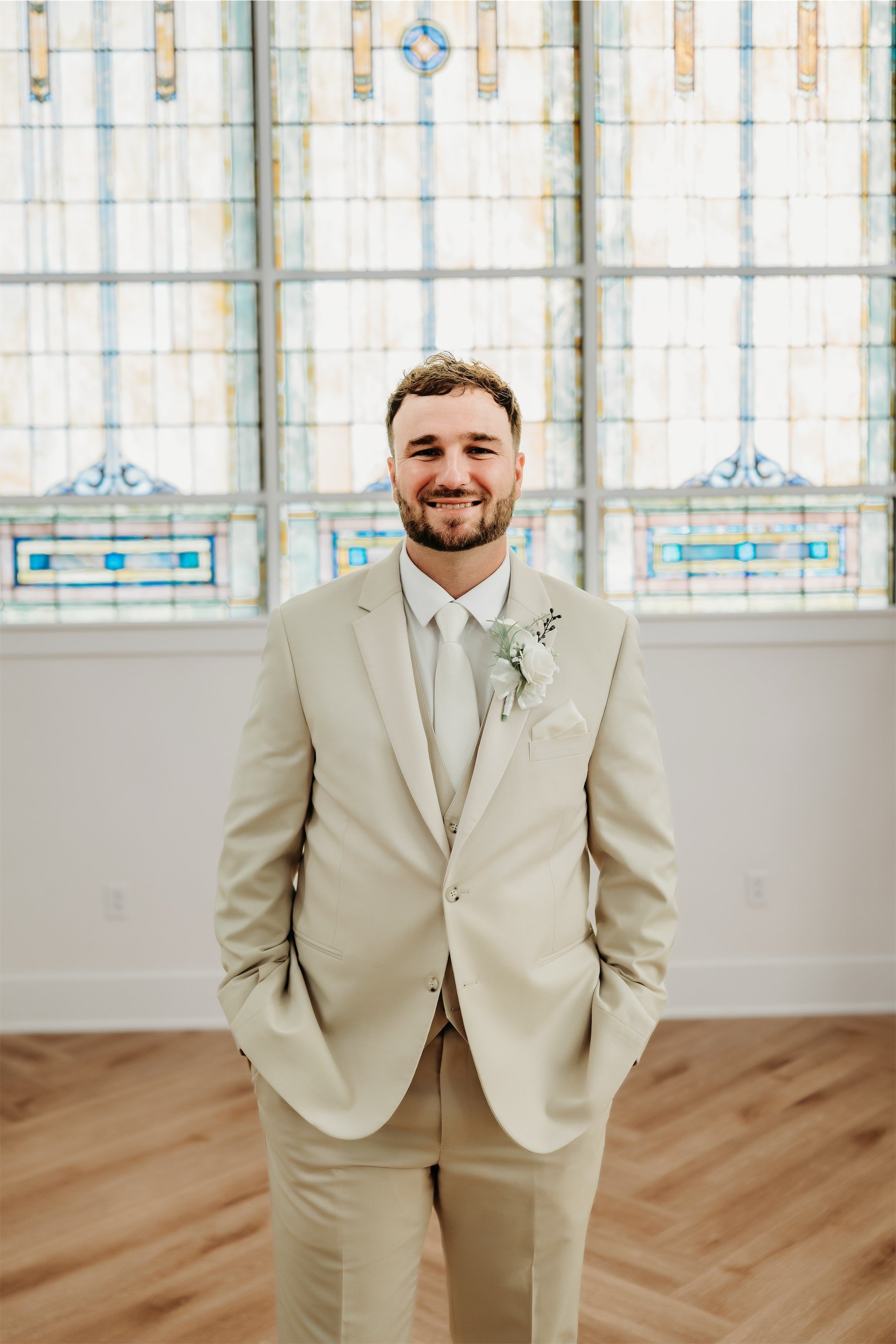 Smiling groom in a tan suit with a sage boutonniere standing in front of stained glass windows
