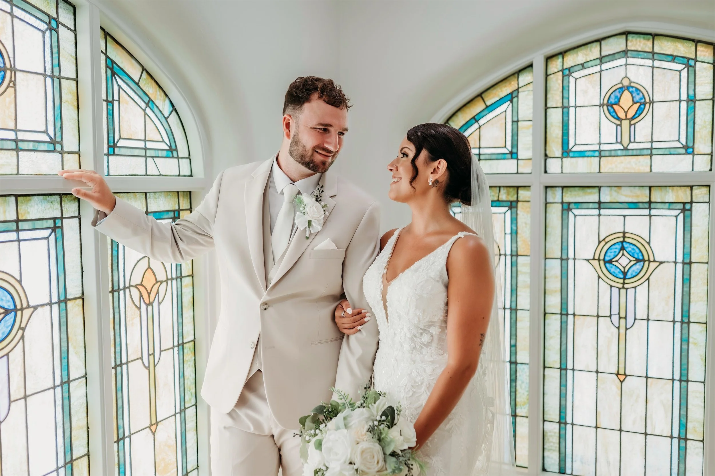 Bride and groom smiling at each other beside arched teal stained glass windows with a white bouquet