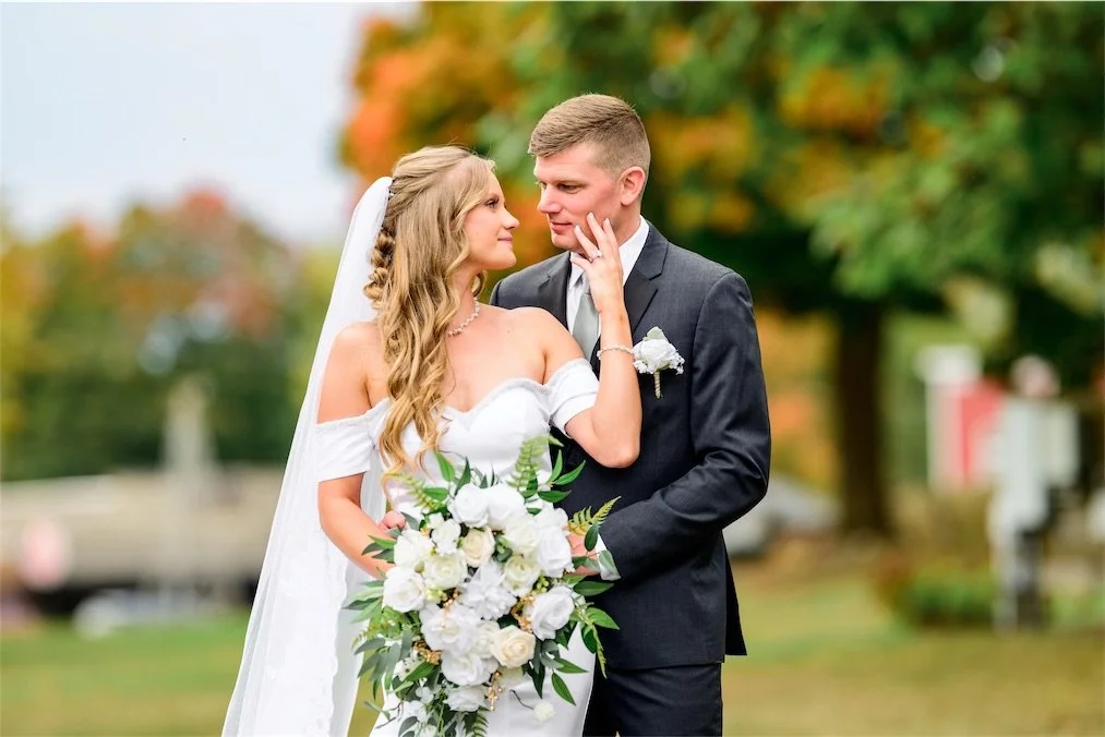 Groom looks at his bride as she touches his face, holding a white bouquet among fall leaves