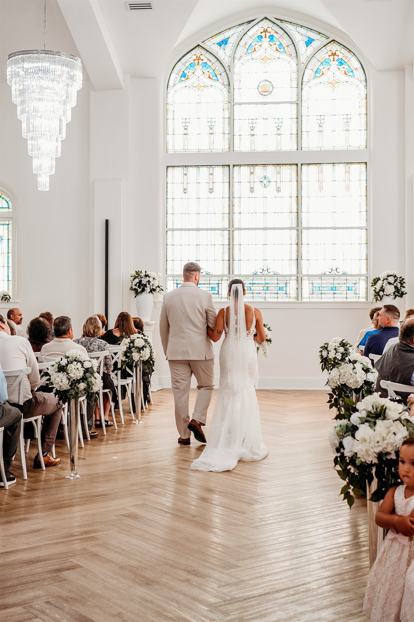 Father walking the bride down a herringbone floor aisle toward a large arched stained glass window