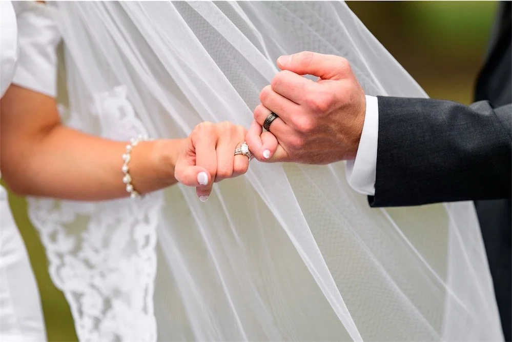 Bride and groom clasp hands with wedding bands showing beneath a sheer lace veil