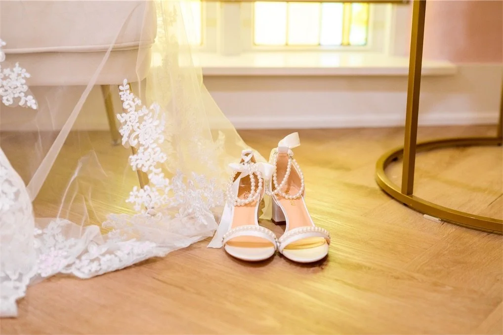 Pearl strap bridal heels on a wood floor beside the lace edge of a wedding veil