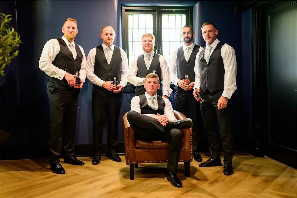 Five groomsmen in black vests pose in a dark navy room, one seated in a leather chair