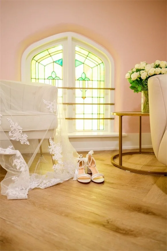 Bridal suite with blush walls, an arched stained glass window, lace veil and heels on the floor