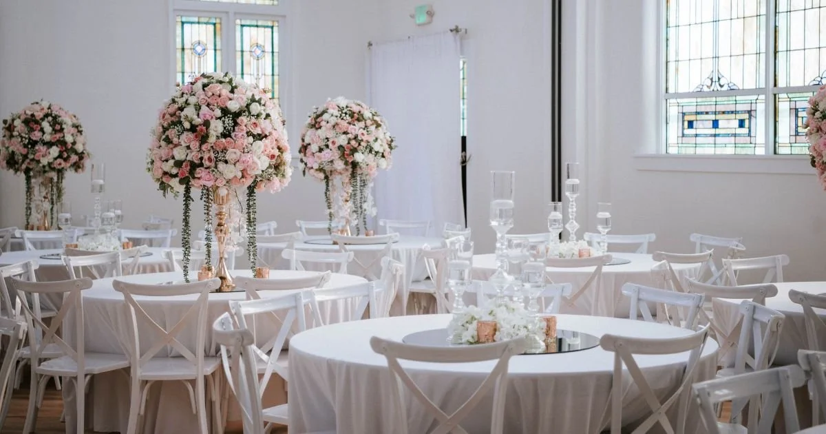 Blush and white floral centerpieces fill a Kaleidoscope Weddings summer reception room with stained glass windows