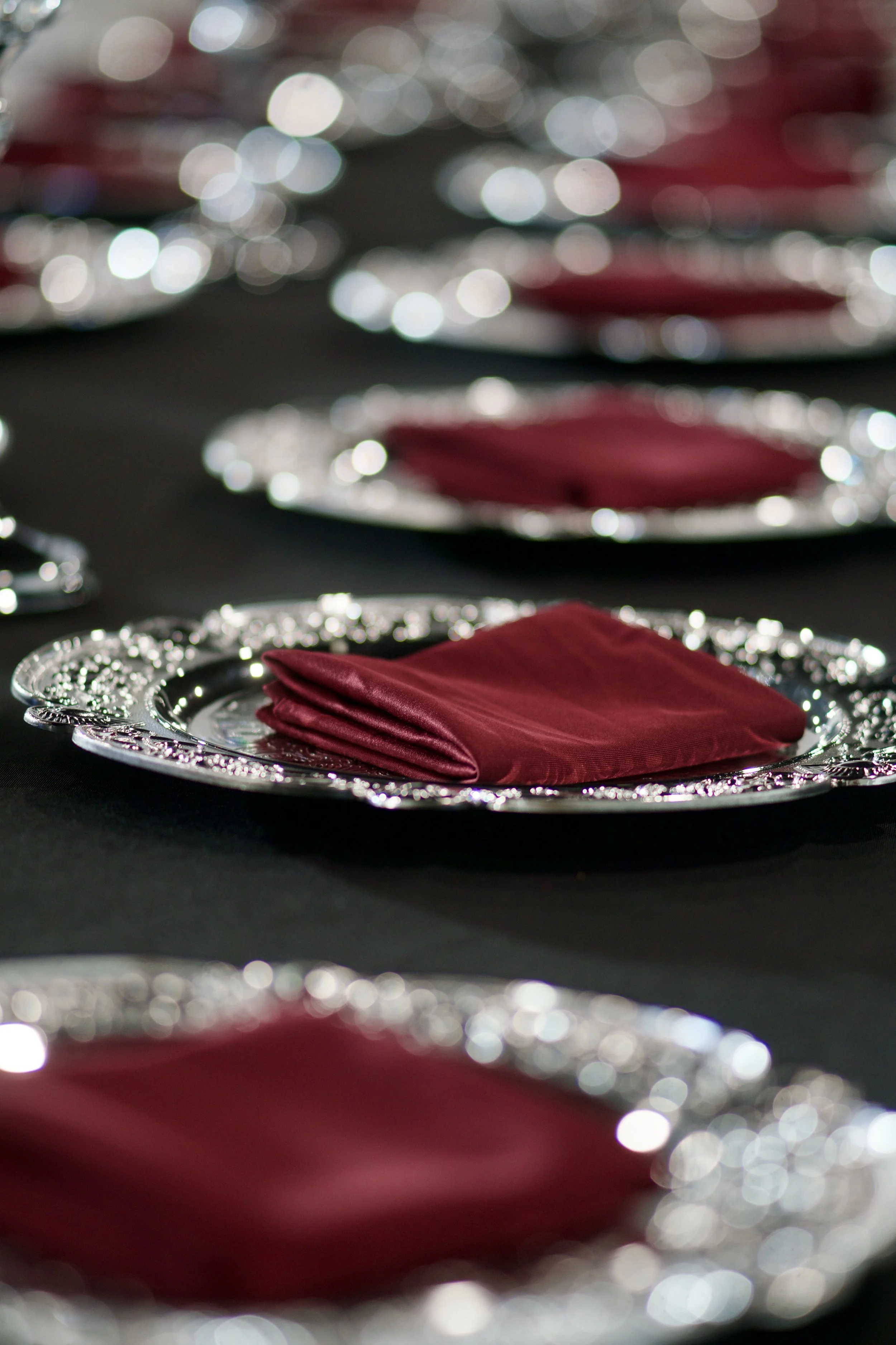 place settings with silver chargers and burgundy napkins down a black linen table