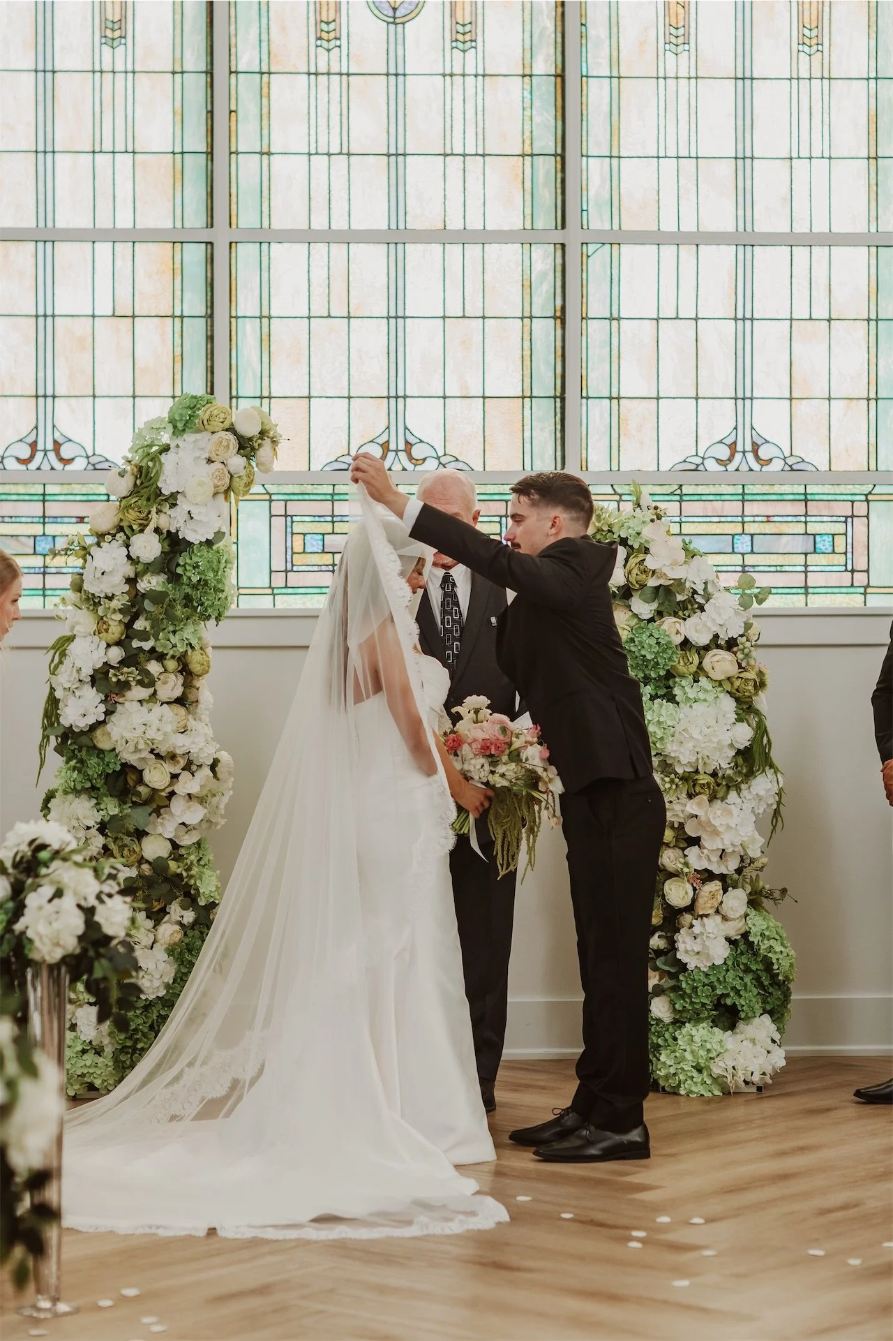 Groom lifting brides cathedral veil at altar before stained glass window
