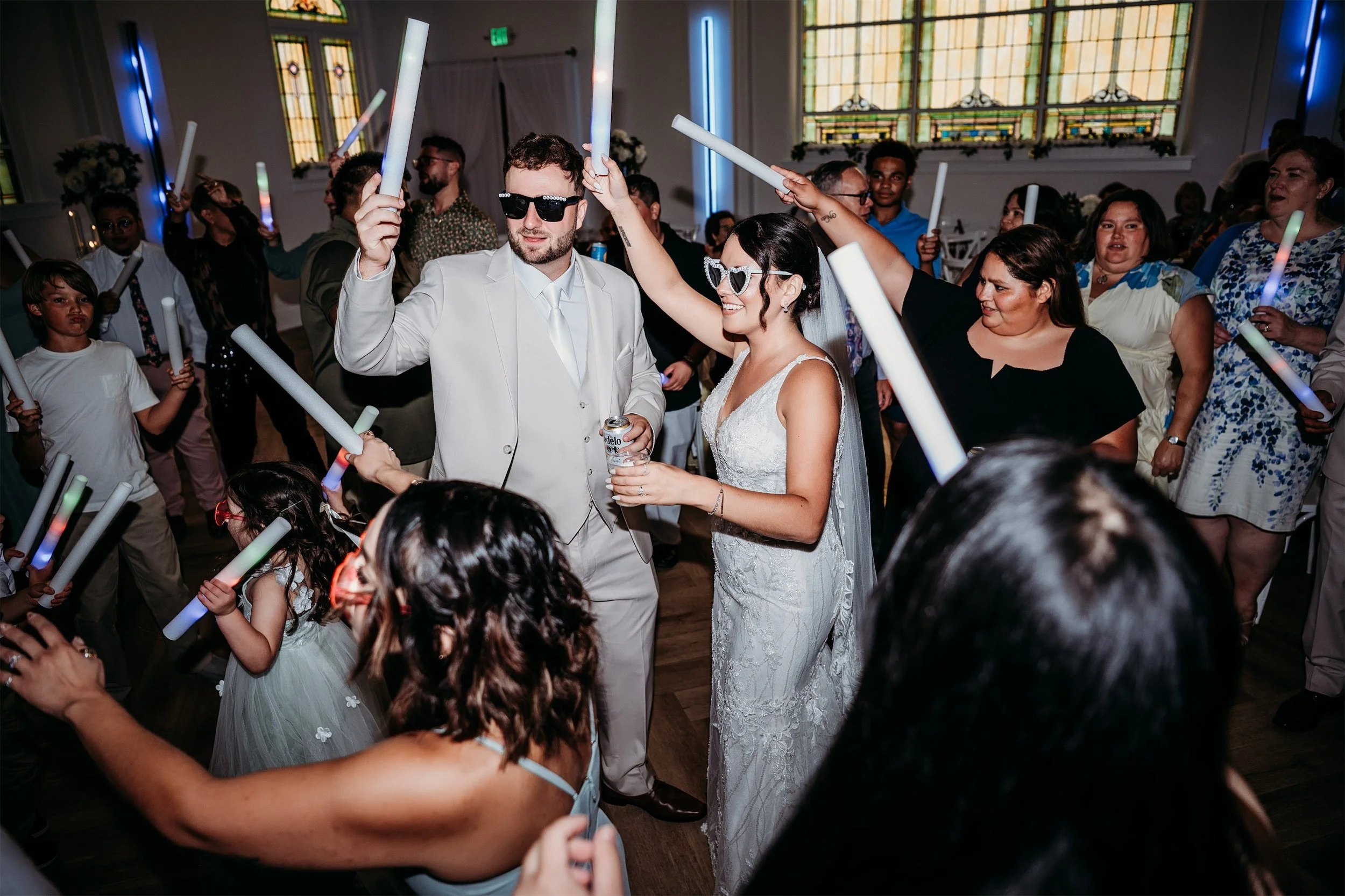 Bride and groom in sunglasses dancing with guests holding glow sticks on the reception dance floor