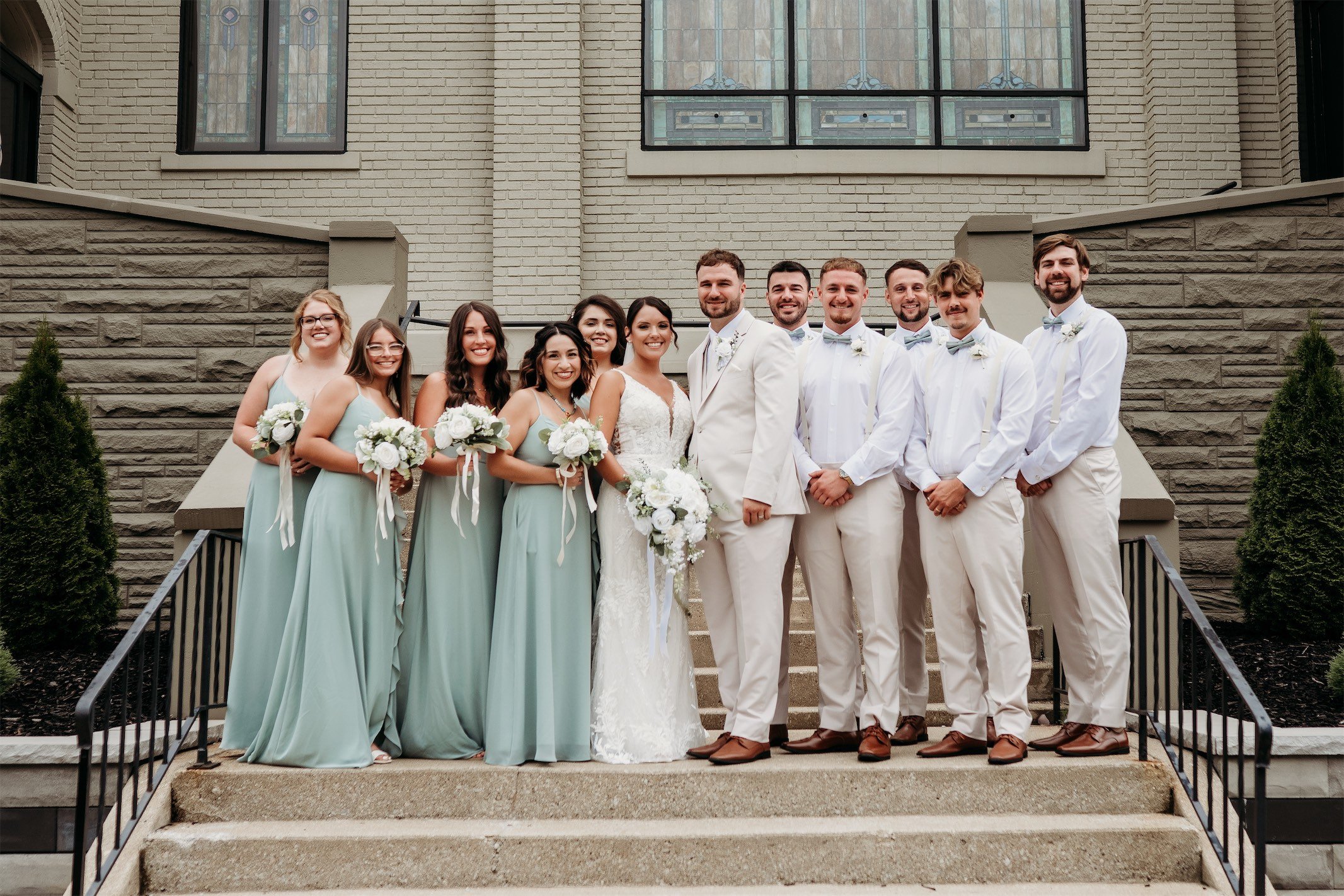 Bridal party of bridesmaids in sage green dresses and groomsmen in tan suits posed on brick church steps