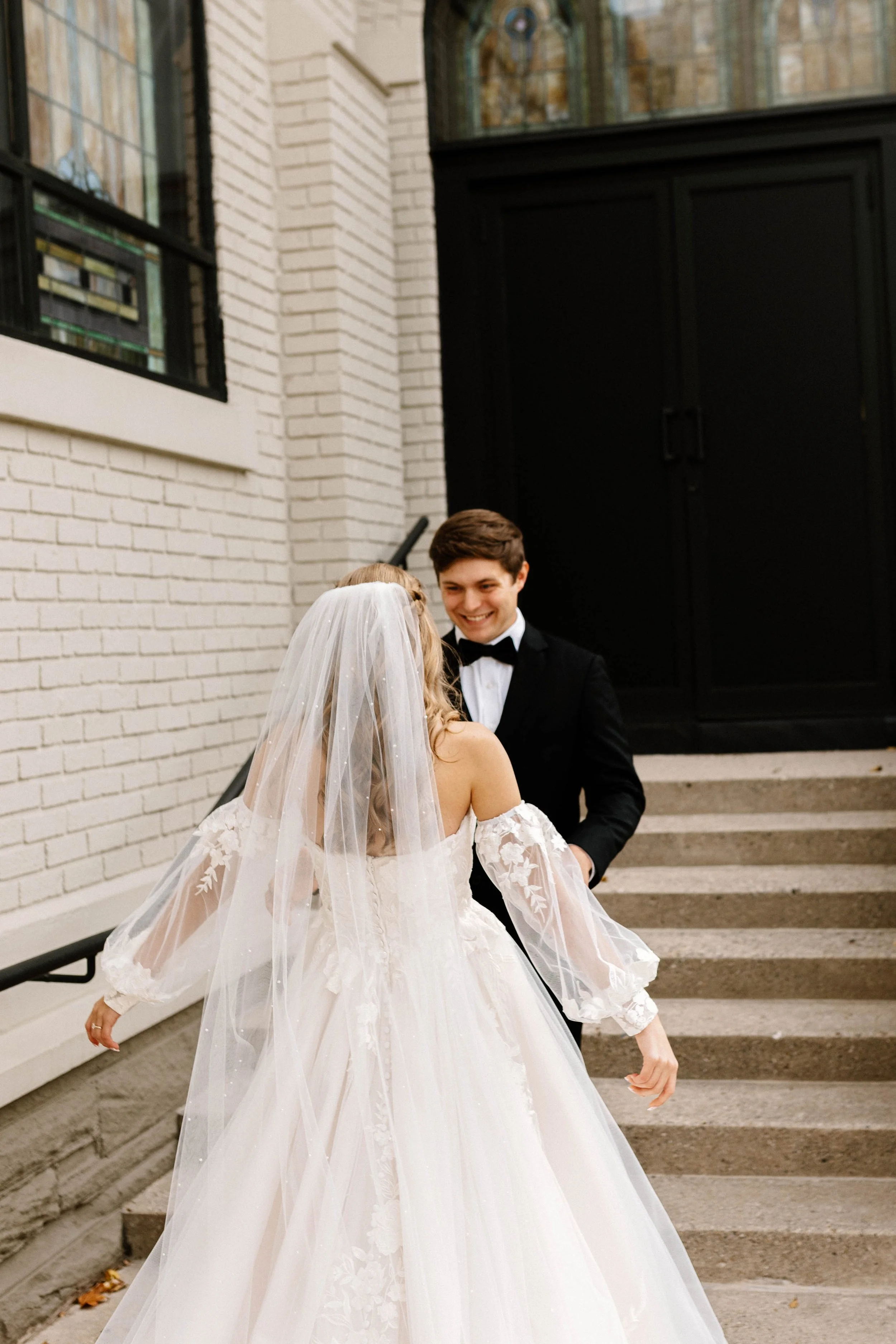 A bride in a white wedding dress and veil dancing with a groom in a black tuxedo outside near a staircase.