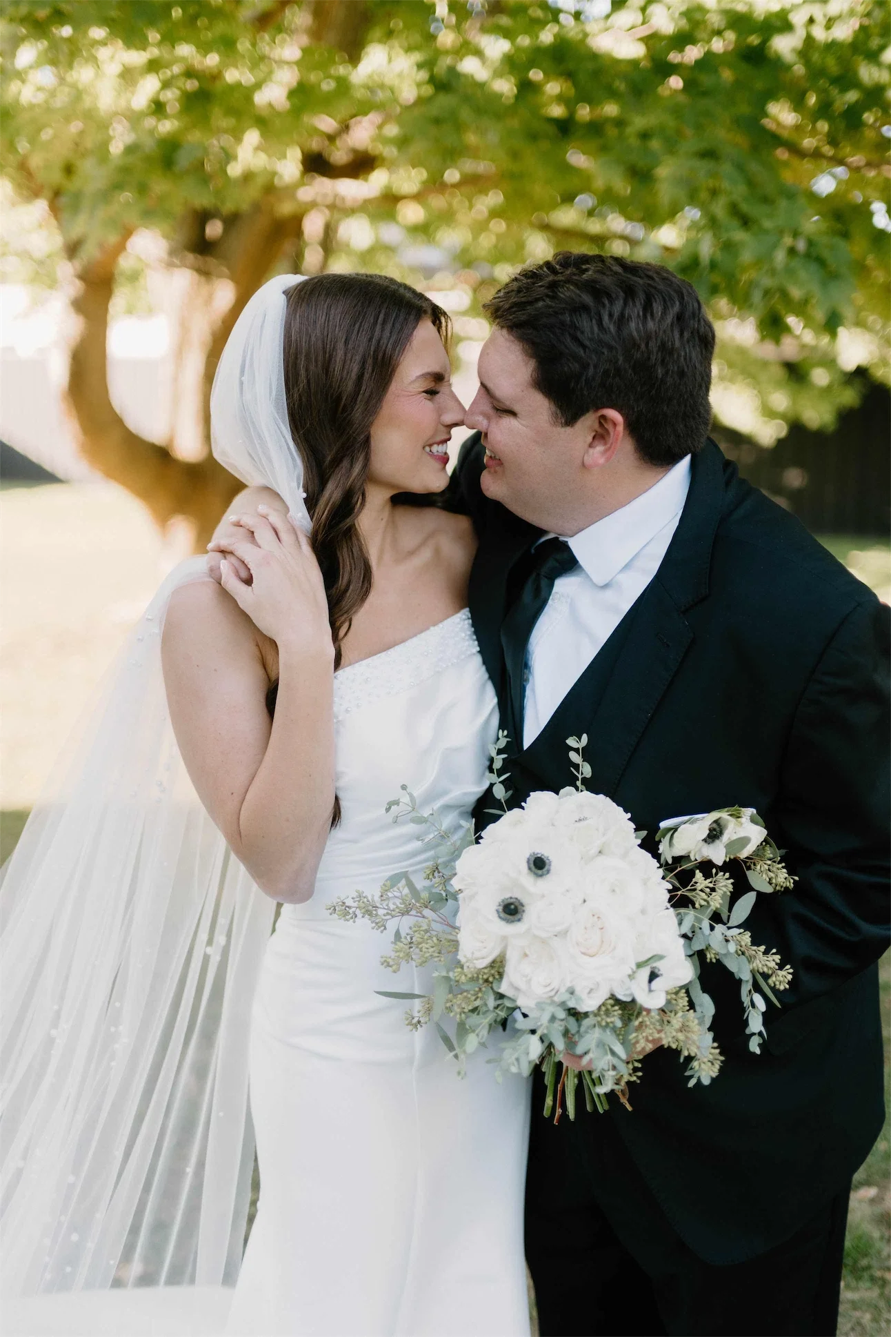 Couple nose to nose under a tree with a white rose and anemone bouquet between them