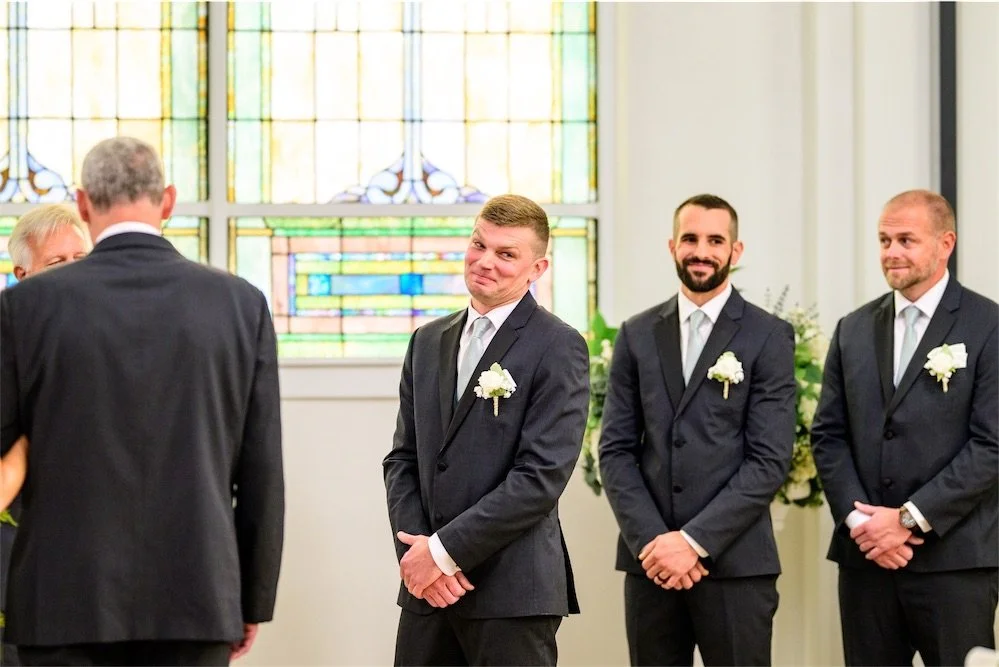Groomsmen with white boutonnieres smile as they watch the ceremony in front of stained glass windows