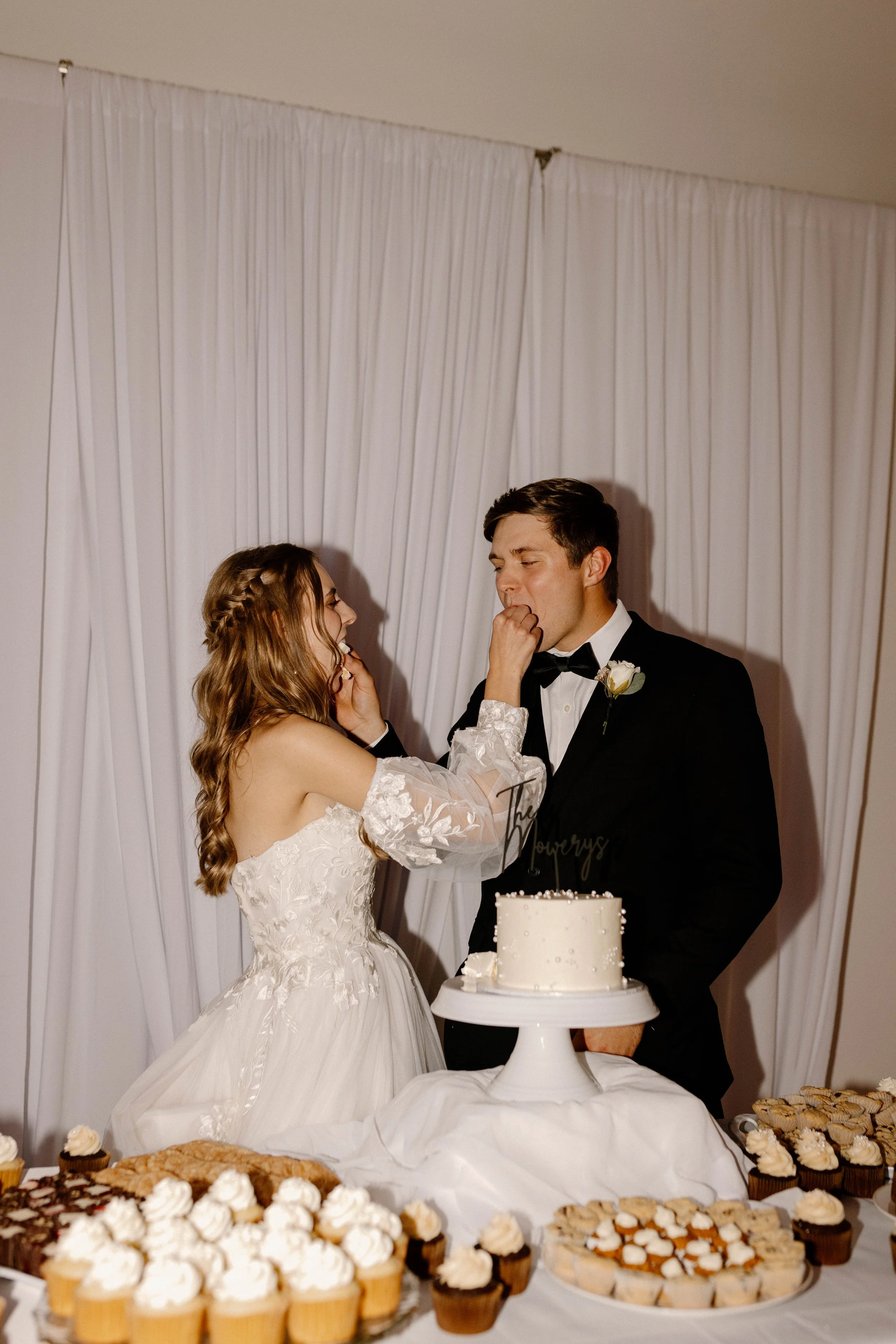 A bride and groom are standing together during their wedding reception, feeding each other cake. The bride is in a white wedding dress with lace details, and the groom is in a black tuxedo with a bow tie. There is a table with a white cake and variou