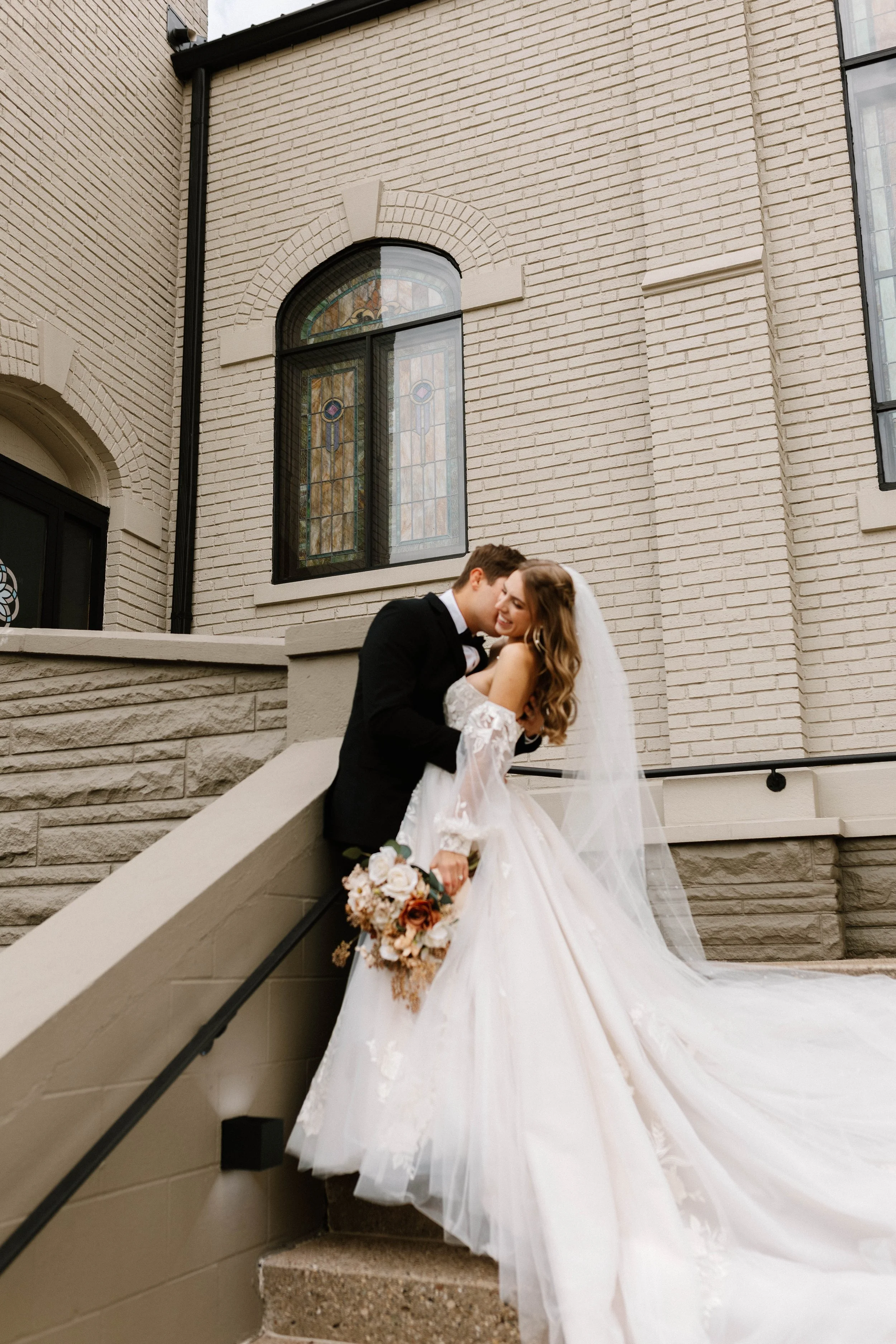 A bride and groom share a kiss on a staircase outside a brick building with stained glass windows, the bride holding a bouquet of flowers.