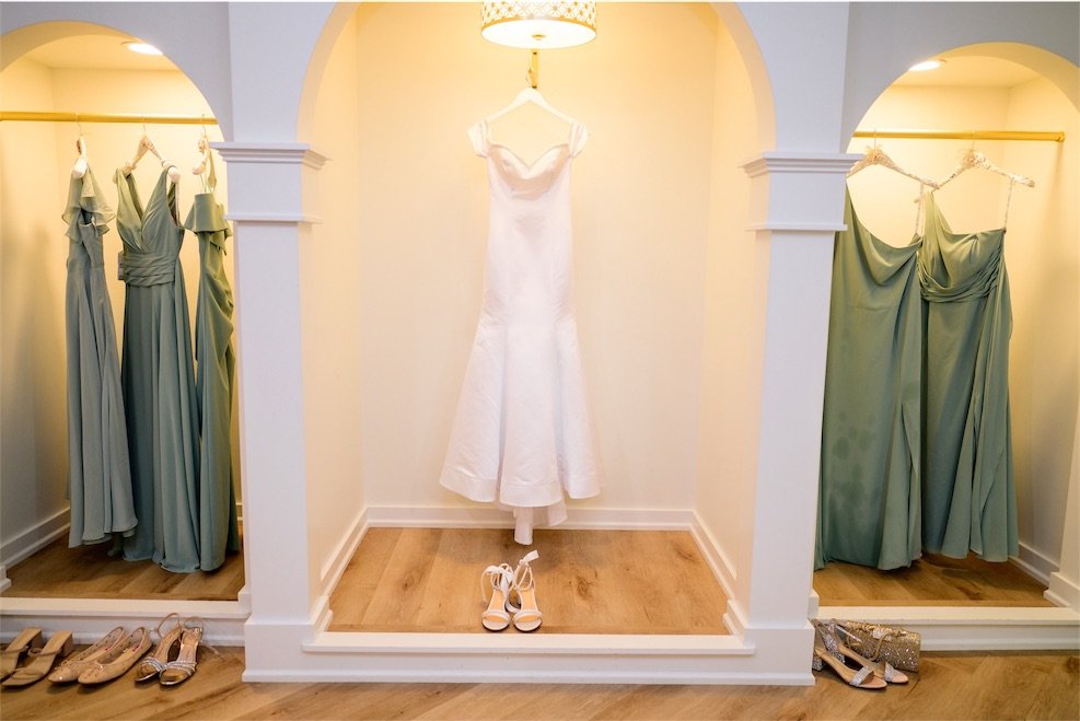 Wedding gown hanging in an arched alcove flanked by sage bridesmaid dresses in the bridal suite