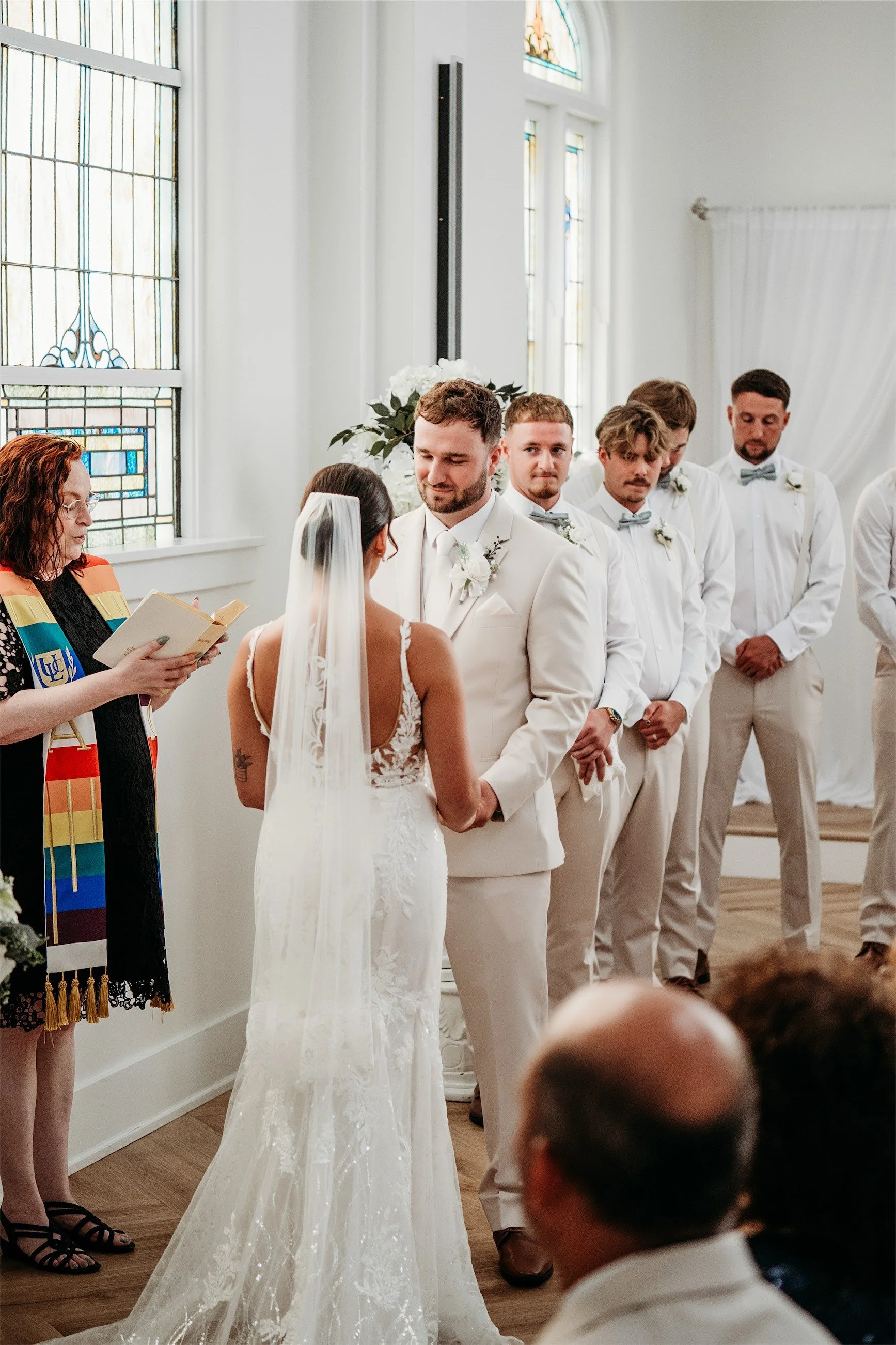 Officiant in a rainbow stole reading to the bride and groom as groomsmen look on during the ceremony