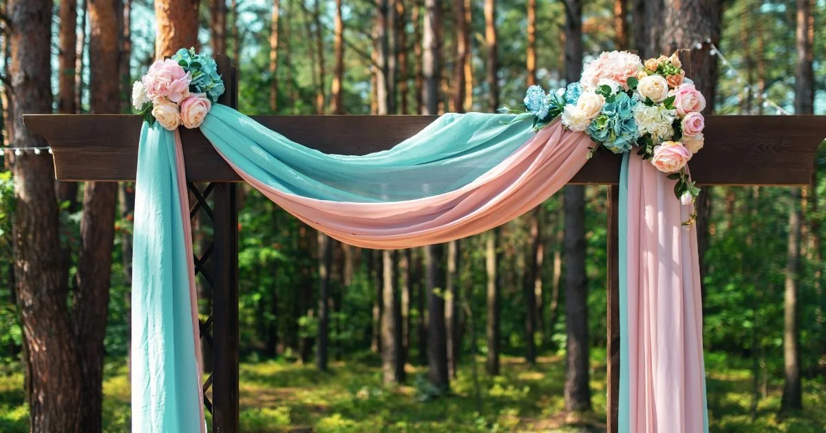 Outdoor summer wedding ceremony arch draped in teal and blush fabric with pink floral arrangements