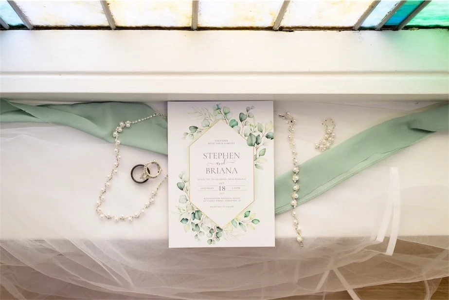 Botanical wedding invitation, rings and pearl necklace arranged on sage green ribbon beside a window