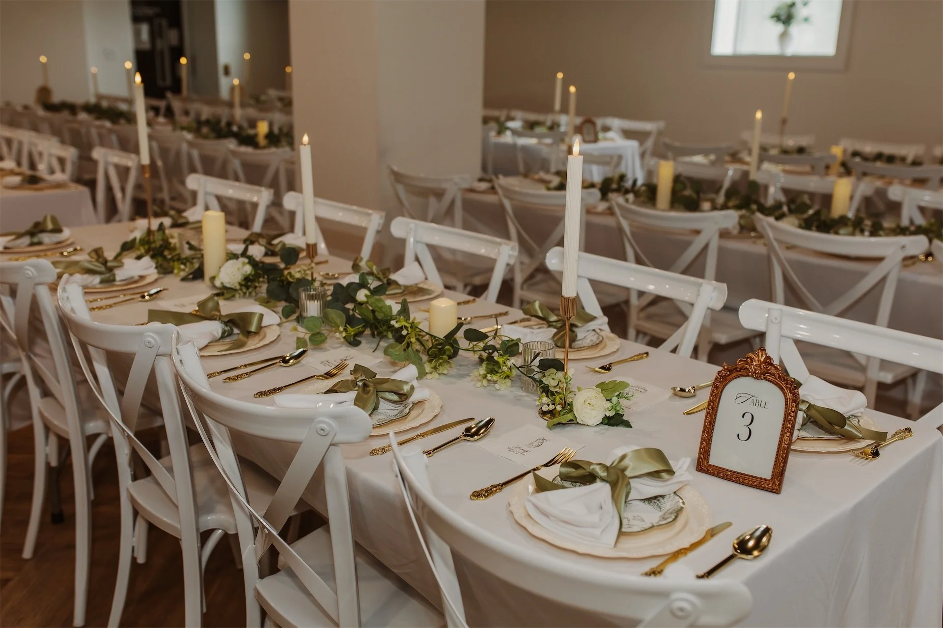 Long reception tables set with gold flatware sage ribbon and taper candles at church wedding venue