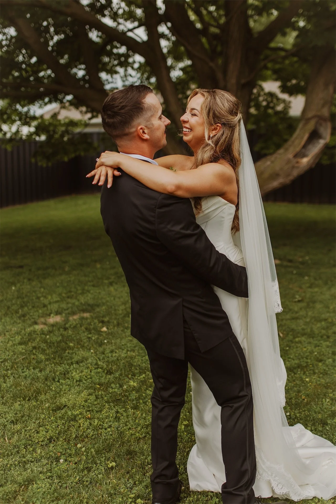 Groom lifting laughing bride off the ground under shade tree on lawn