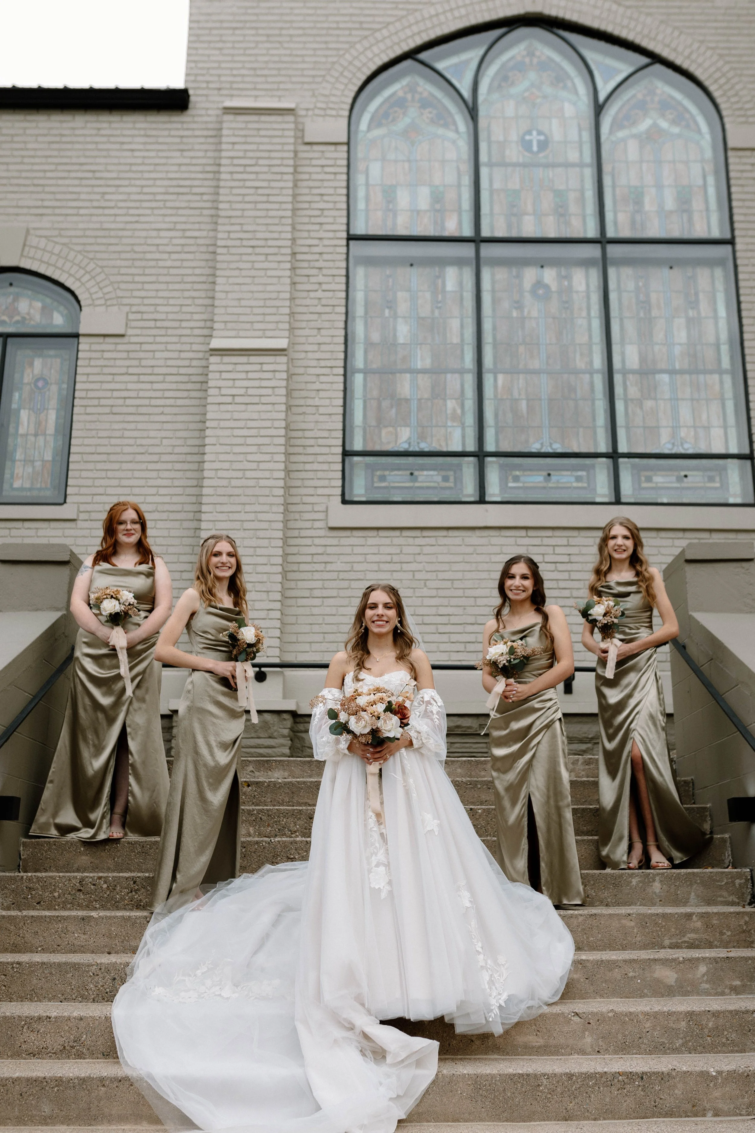 A group of five women, including a bride in a white wedding gown, standing on stairs outside a church with large stained glass windows. The bride holds a bouquet, and four bridesmaids in matching gold-colored dresses hold smaller bouquets.