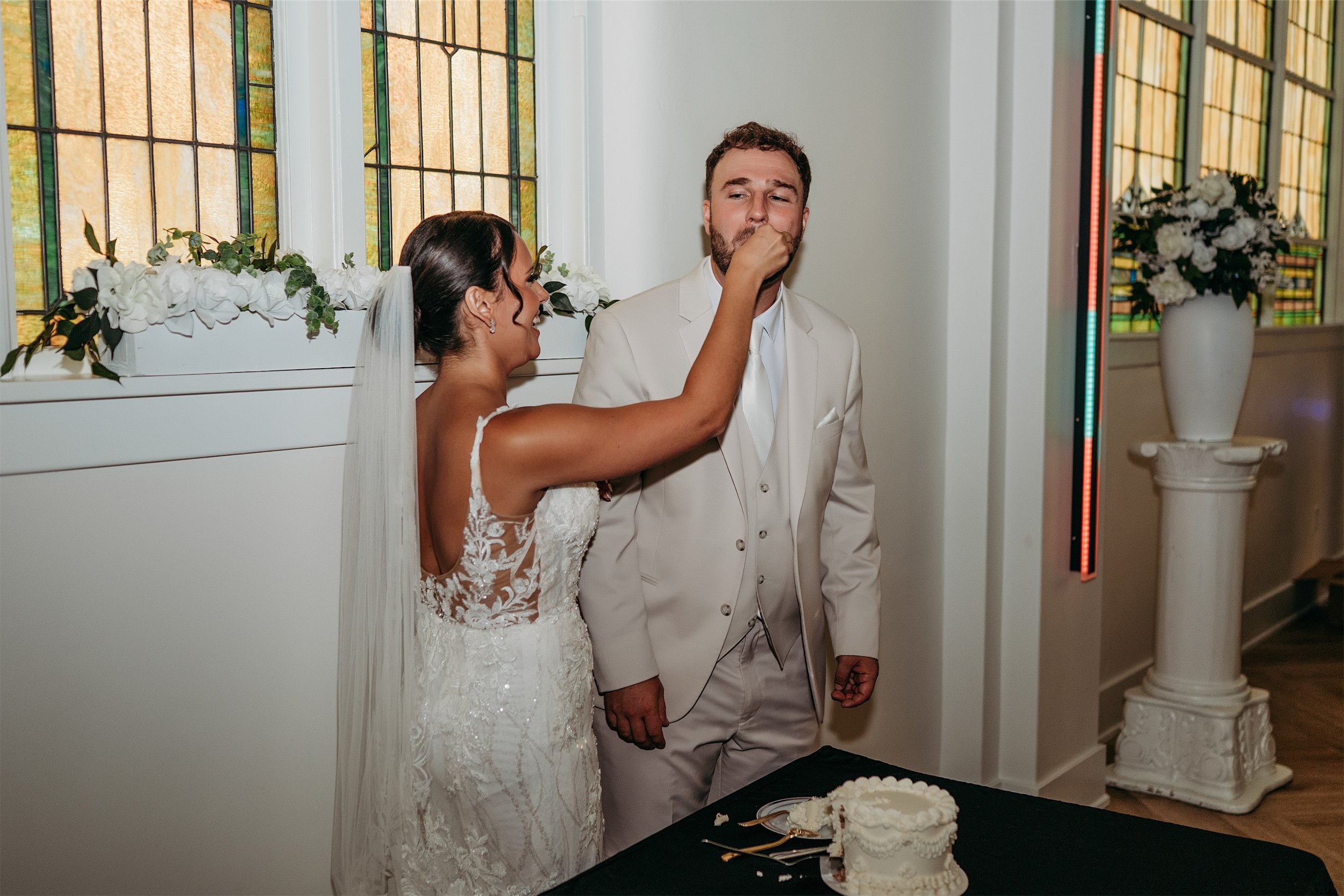 Bride feeding groom a bite of wedding cake beside a white floral pedestal and stained glass windows