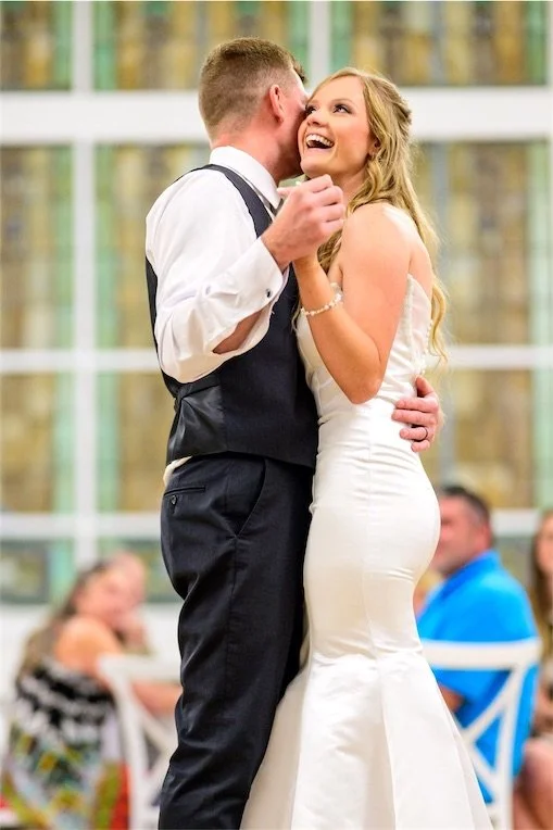 Groom whispers to his laughing bride as they dance at the reception with guests behind them